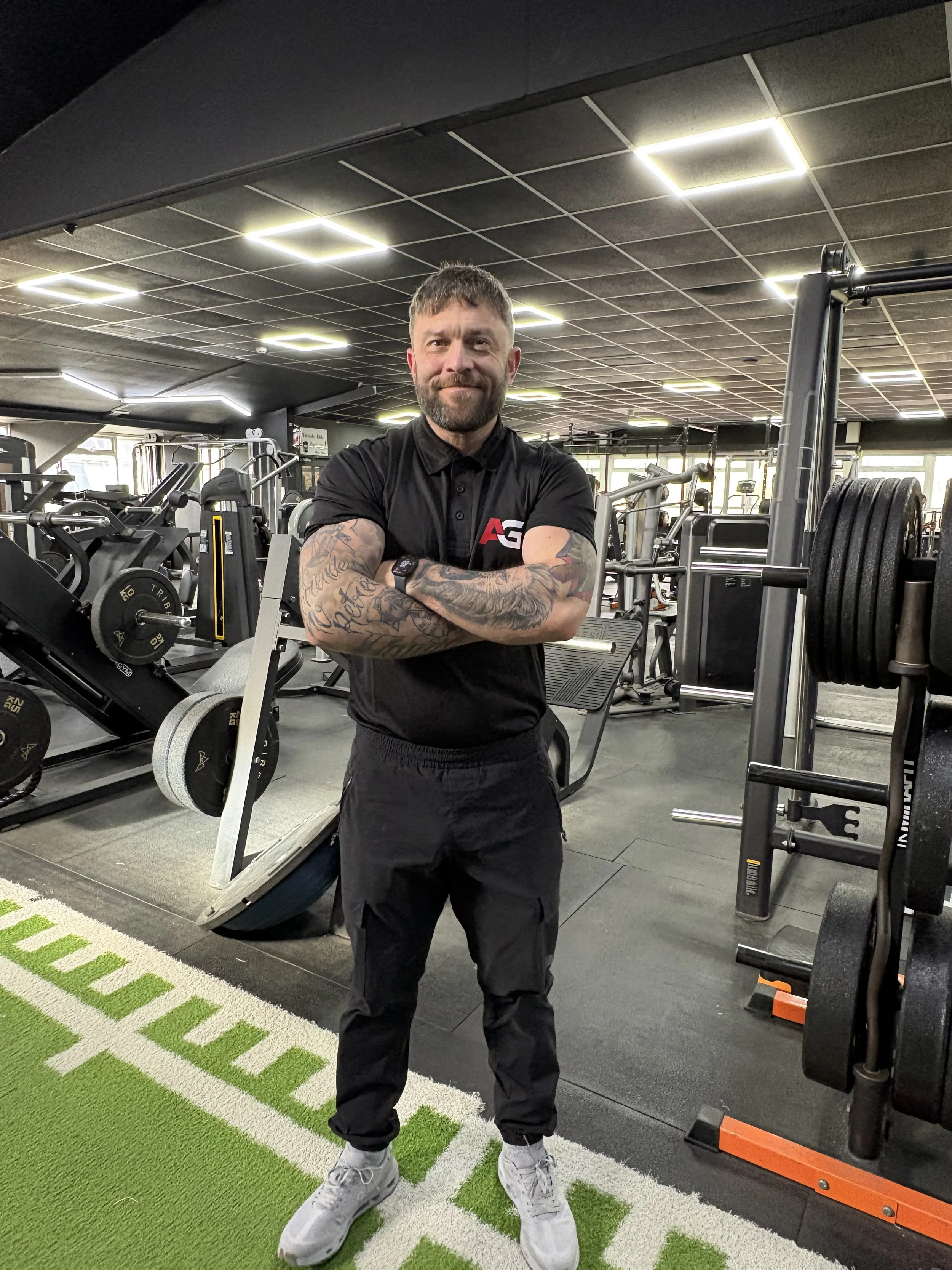 A man with tattoos wearing a black polo and black pants standing with arms crossed in a gym.