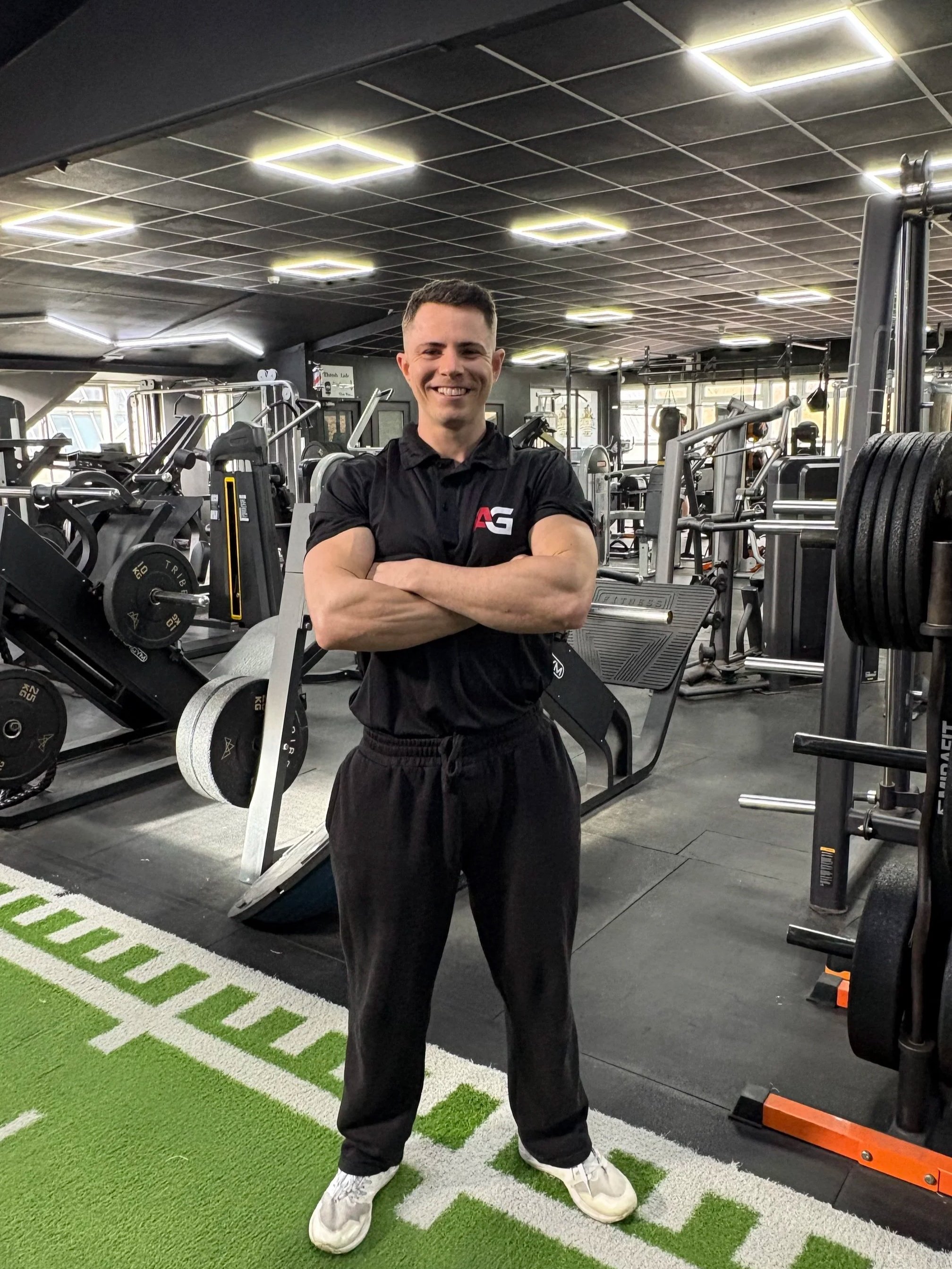 A smiling man in black workout clothes standing with arms crossed in a gym with various exercise equipment in the background.