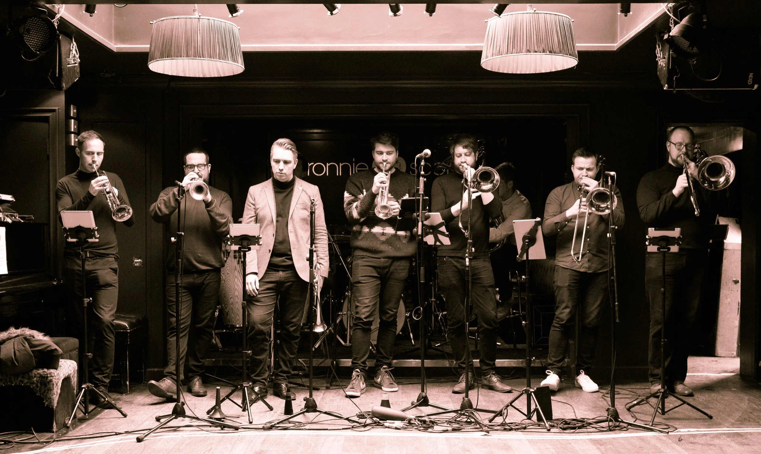 A jazz band performing on stage with eight members playing various instruments, including trumpets and trombones, in a dimly lit venue with black walls and large hanging lamps.