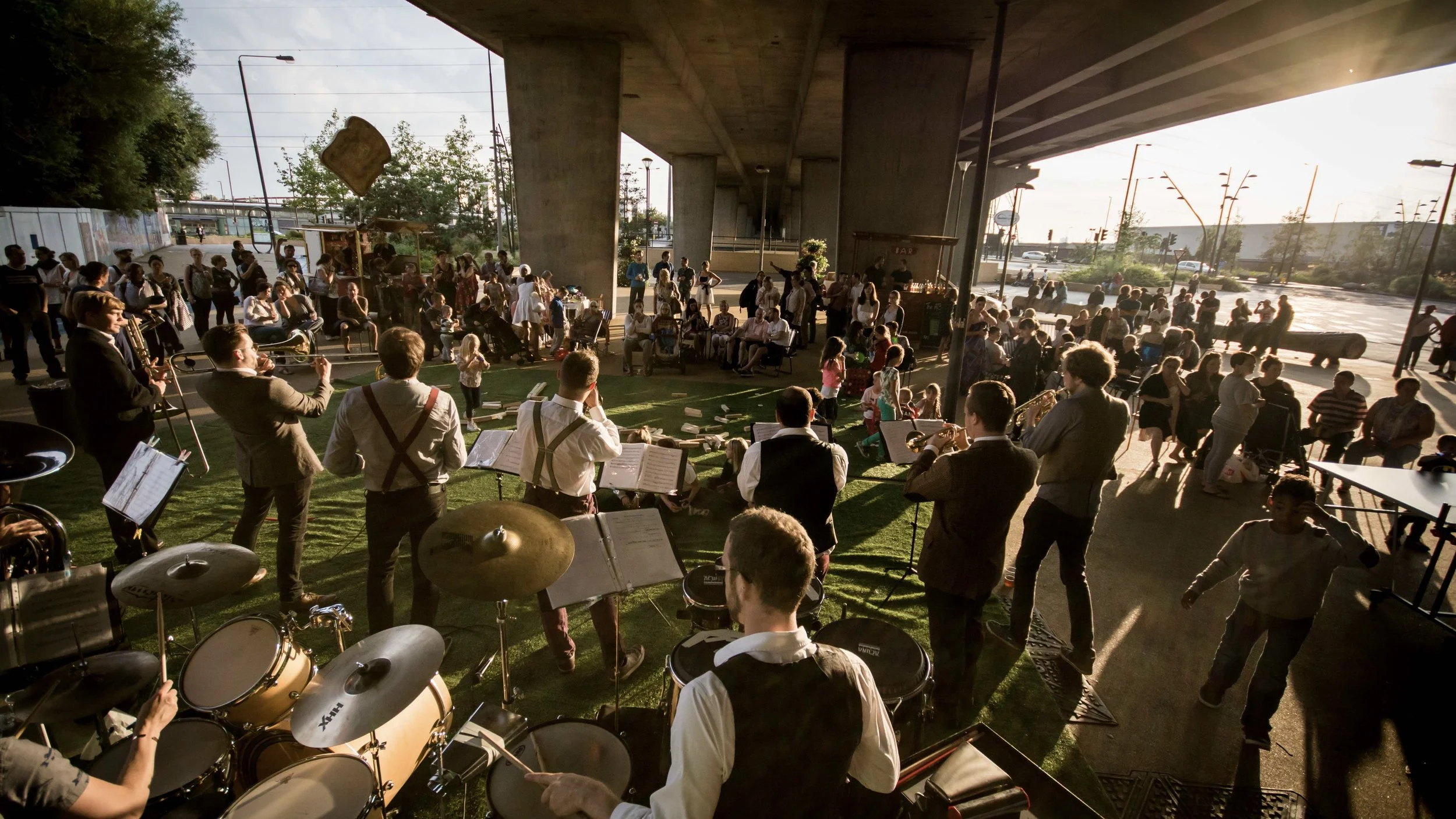 Live jazz band performing under a bridge with an audience enjoying the outdoor event during sunset.