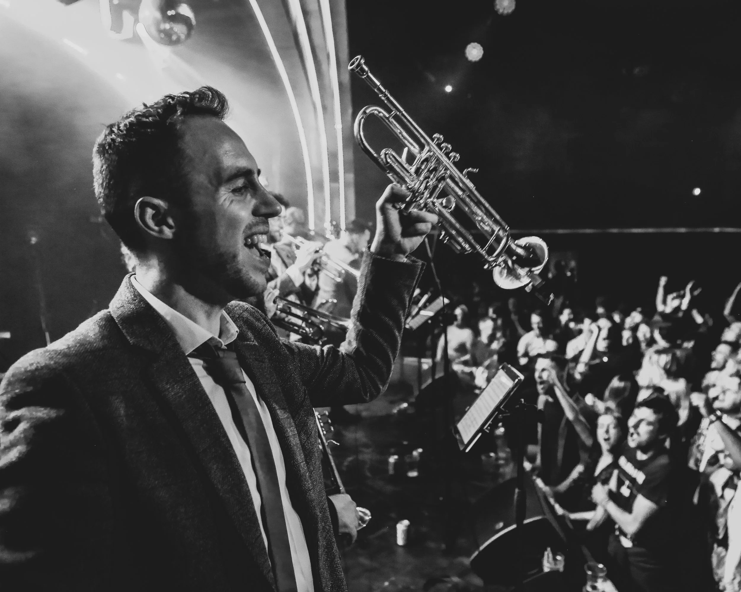 Black and white photo of a man in a suit playing a trumpet at a lively gathering or concert, surrounded by other musicians and an enthusiastic audience.