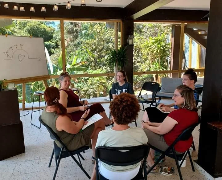Six people sitting in a circle in a sunlit room with large windows showing greenery outside, engaged in discussion. A whiteboard with a drawing of a cloud, a house, and a heart is visible in the background.