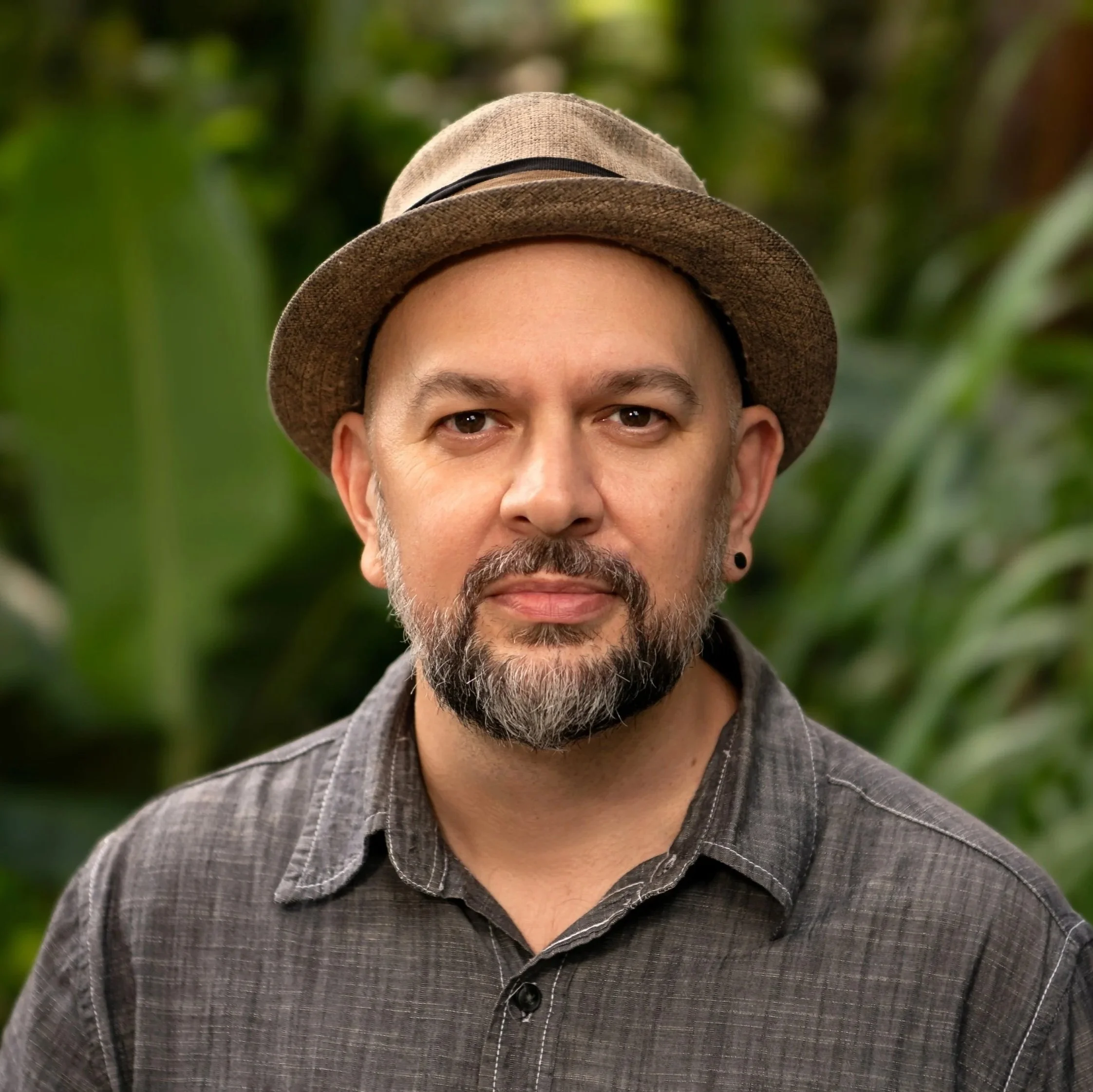 A smiling man with a beard and mustache, wearing a brown cap and black shirt, standing in front of a stone wall.