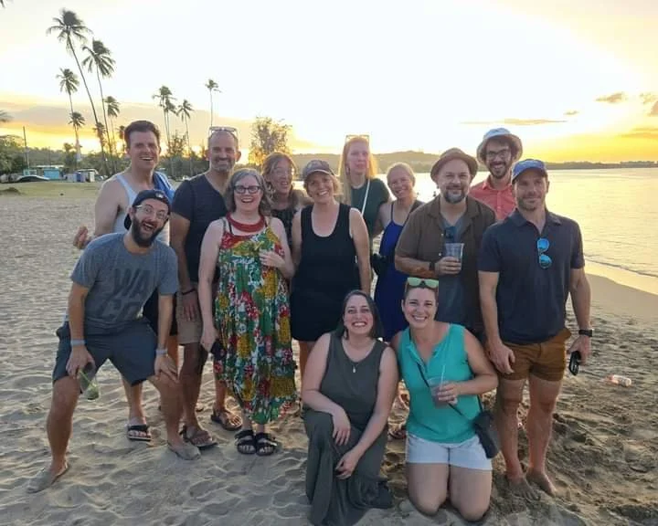 Group of people enjoying a beach sunset, some holding drinks, in casual summer clothes, with palm trees in the background.