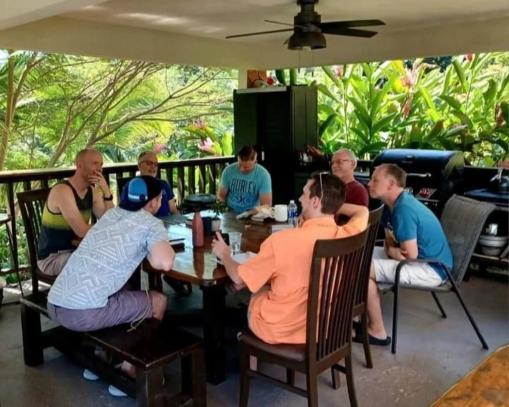 Group of seven men sitting around a wooden outdoor dining table on a porch with lush greenery in the background.