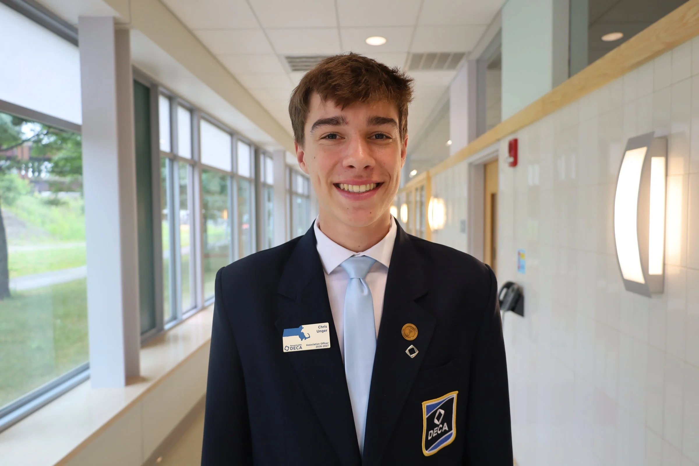 Young man smiling in a hallway wearing a formal jacket with DECA and school badges.
