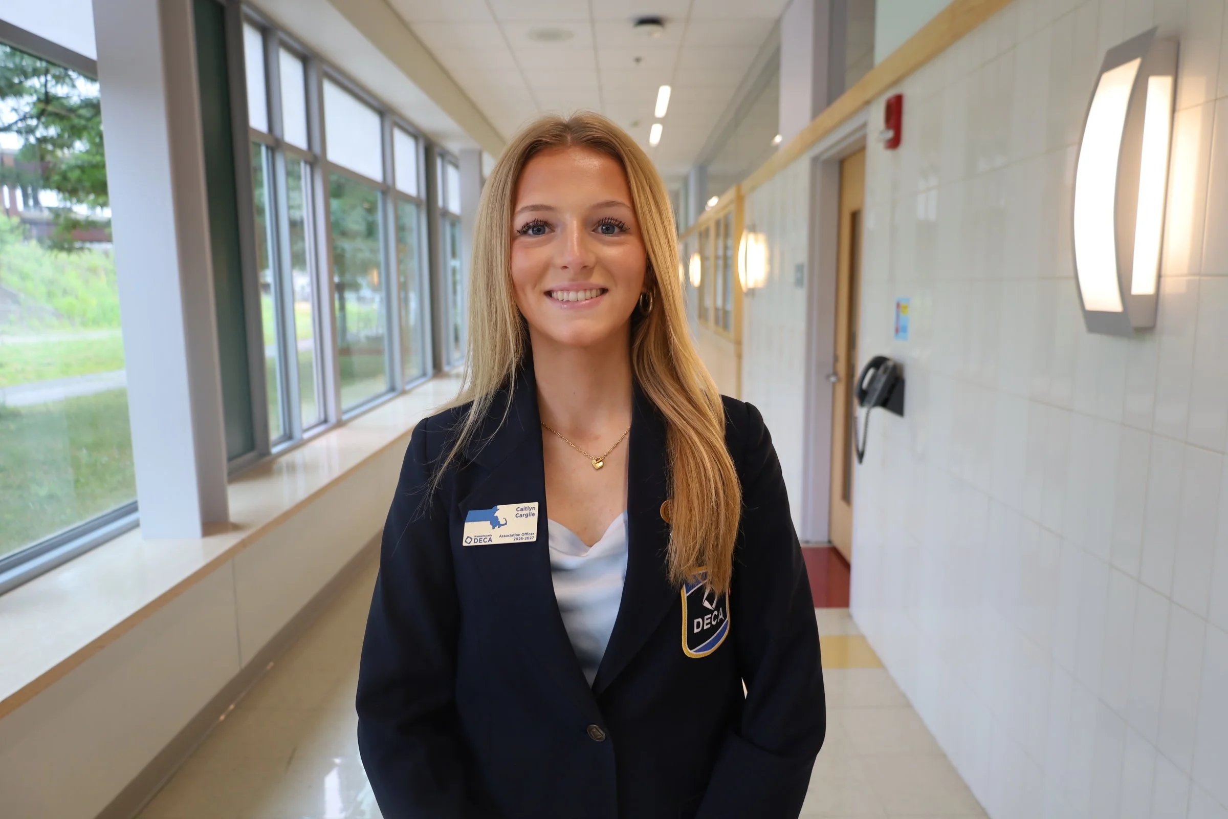 A young woman with long blonde hair wearing a black blazer and a name tag standing in a hallway with large windows and modern lighting.