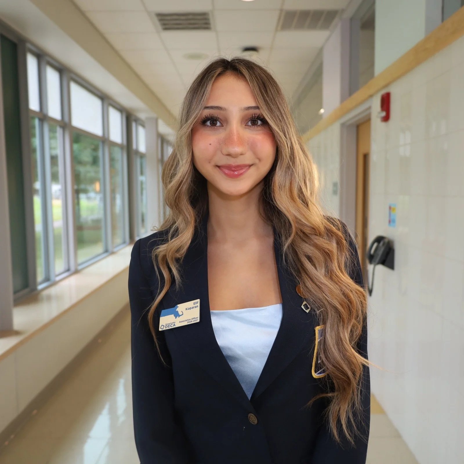 A young woman in professional attire standing in a corridor, smiling at the camera, wearing a name badge with her name and association logo.