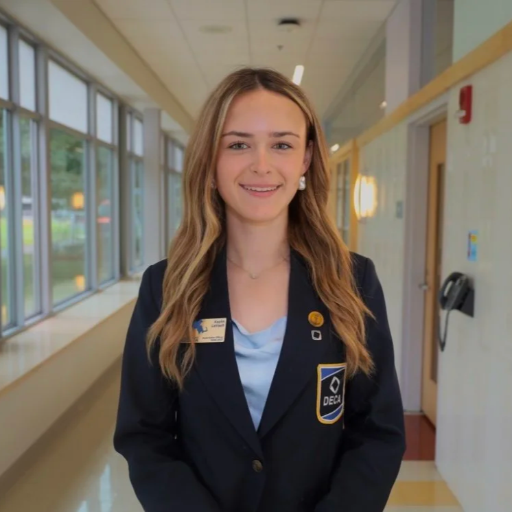 Young woman in DECA honor society blazer standing in school hallway with windows and yellow walls.