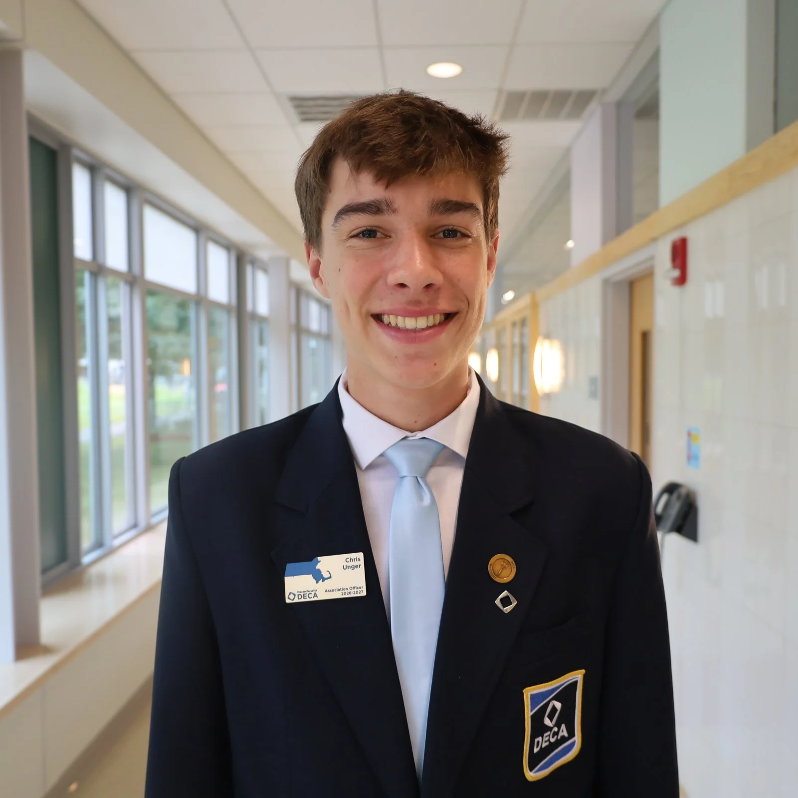 Young man smiling, dressed in a black DECA blazer with patches and name tag, standing in a corridor with large windows.