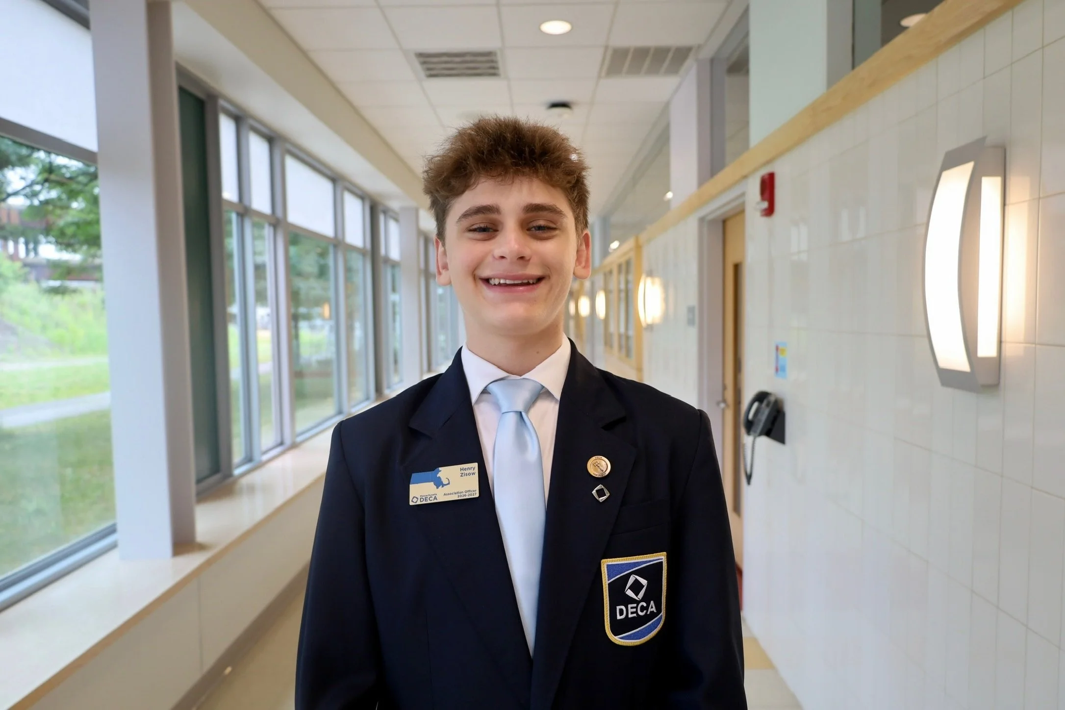 Young man wearing a DECA badge and blazer, standing in a building corridor with large windows and natural light, smiling at the camera.