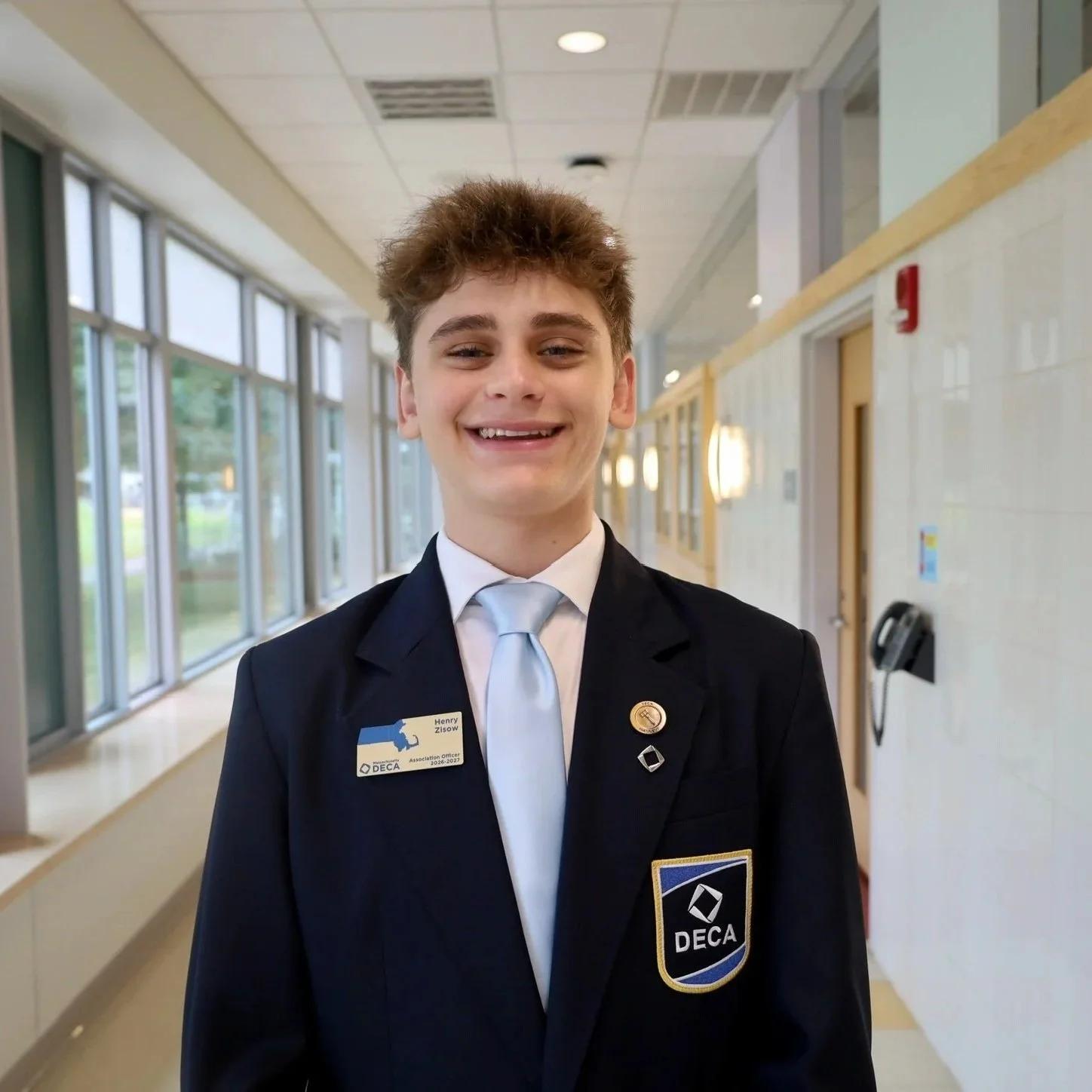 Young man in formal suit smiling, standing in school hallway with large windows, wearing a DECA badge and pins.