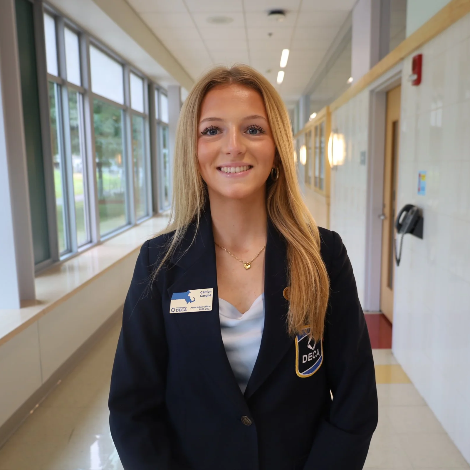 A young woman with long blonde hair, wearing a black blazer with a Name tag, standing in a brightly lit indoor area near large windows and a hallway.