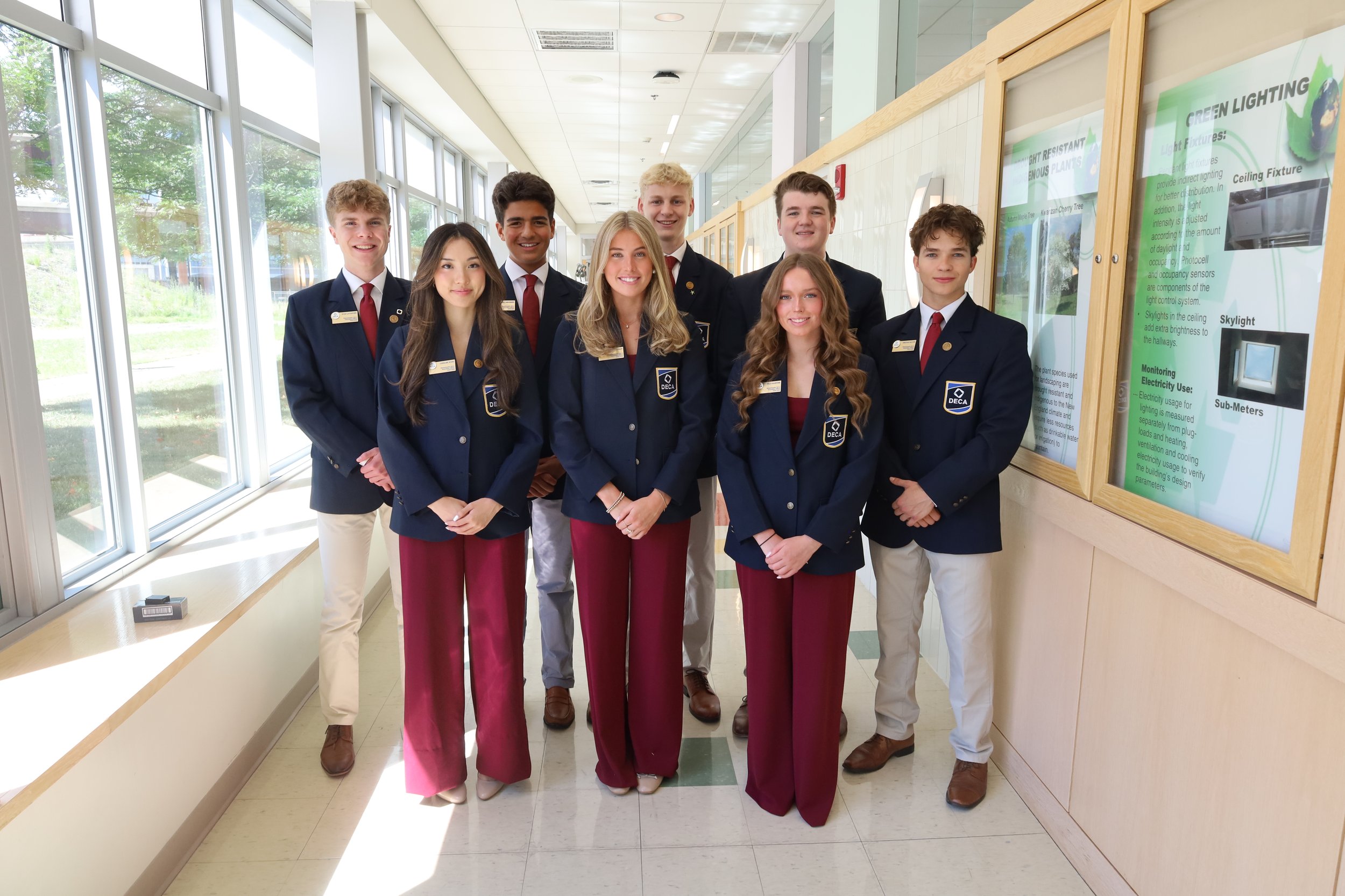 A group of nine young people wearing navy blazers with school or club insignias, standing in a brightly lit hallway with large windows, smiling at the camera.