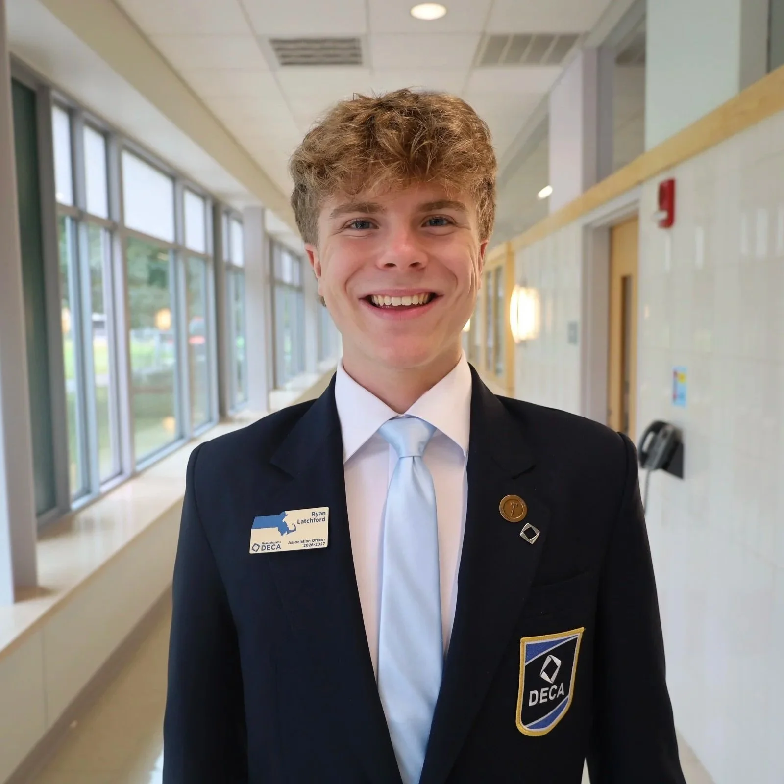 Young man wearing a black uniform with an 'NCIA DECA' badge and pins, smiling in a hallway with large windows.