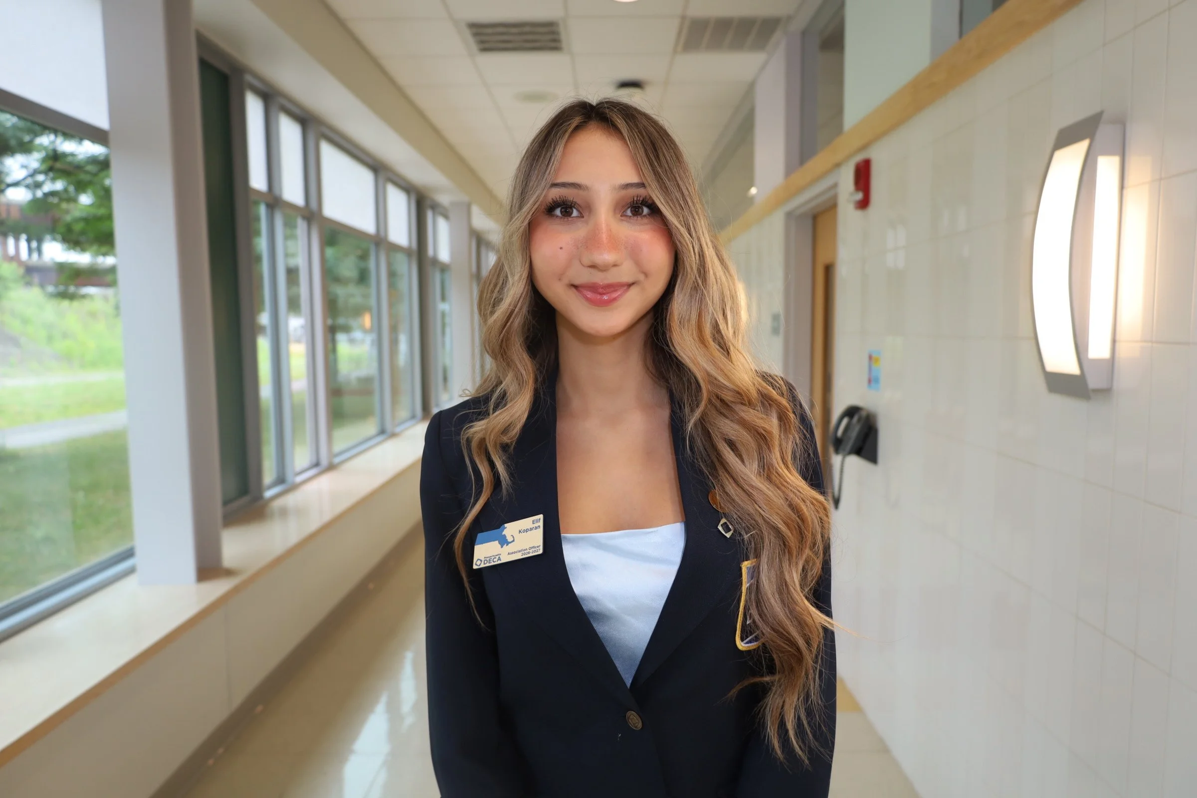 A young woman with long, wavy blonde hair and light skin, wearing a dark blazer with a name tag, smiling inside a building with large windows and white walls.