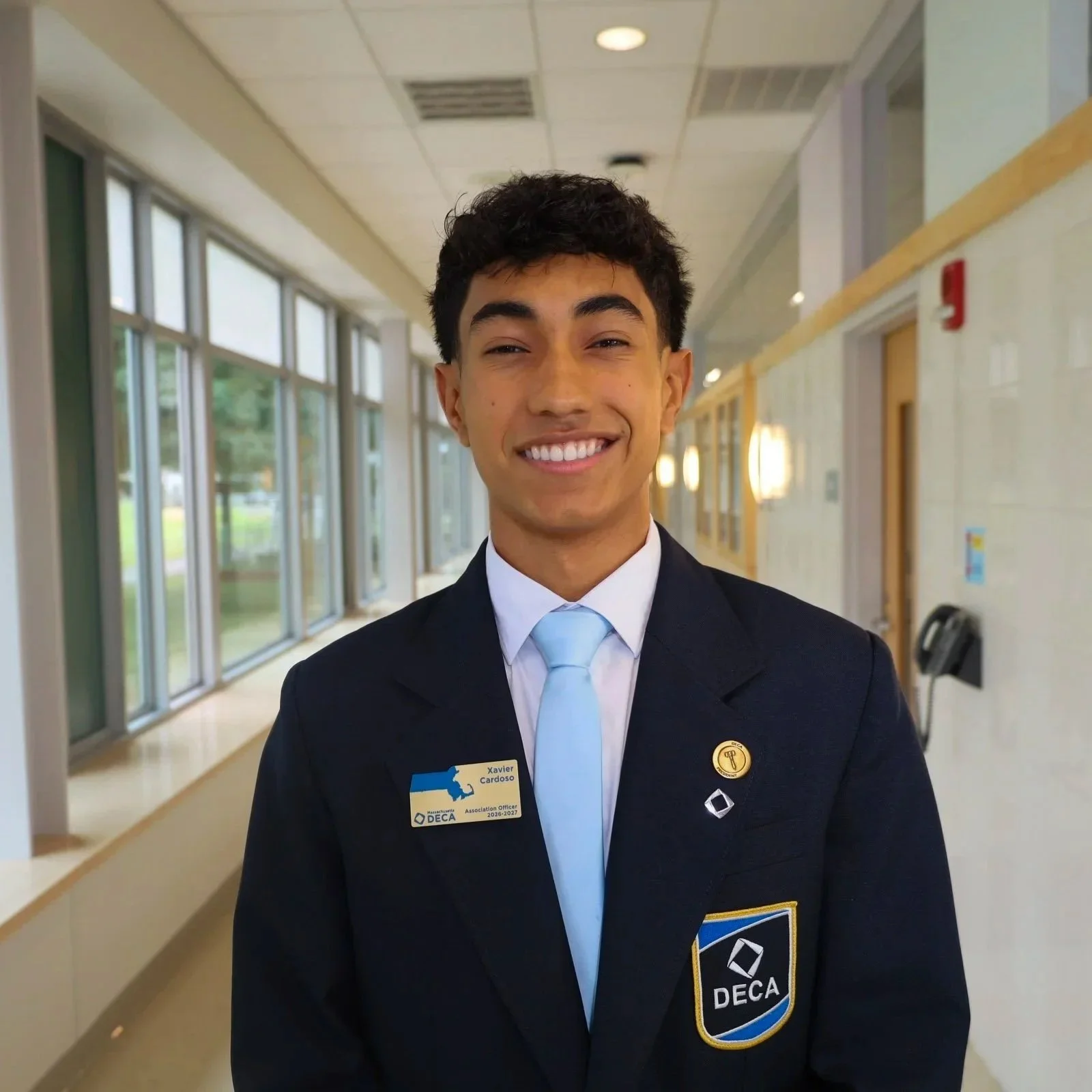 Young man in a professional uniform smiling in a hallway, wearing a DECA badge and pins.