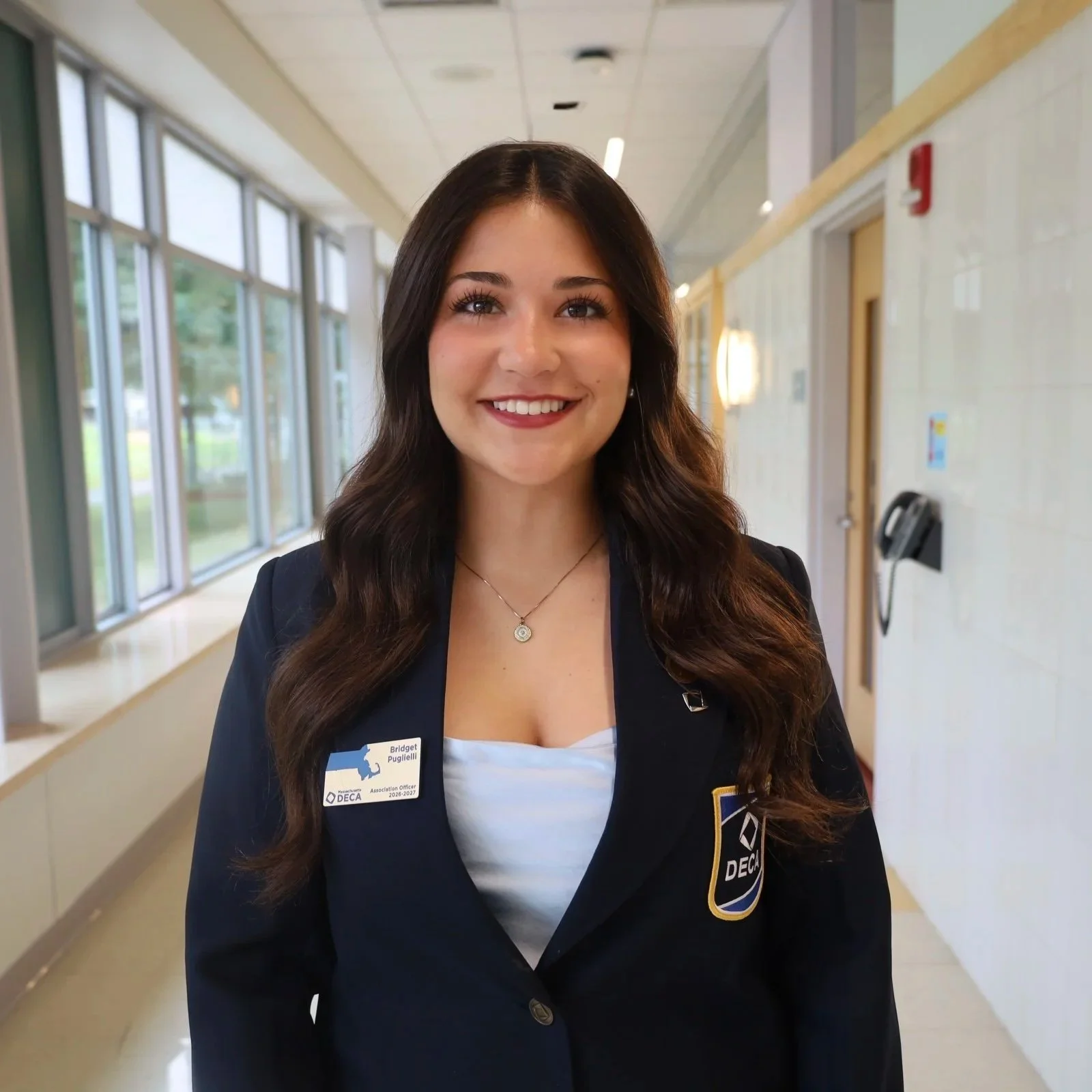 A young woman with long, dark, wavy hair, smiling, wearing a name tag, and a navy blazer with a DECA patch, standing in a hallway with large windows on the left and a door on the right.