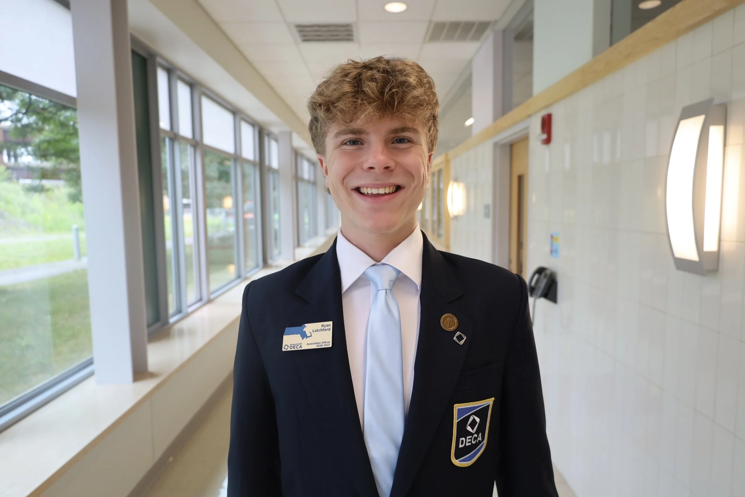 A young man with light brown curly hair, wearing a dark blazer with badges, a white shirt, and a light blue tie, smiling in a brightly lit indoor space with large windows, a beige tiled wall, and mirrors.