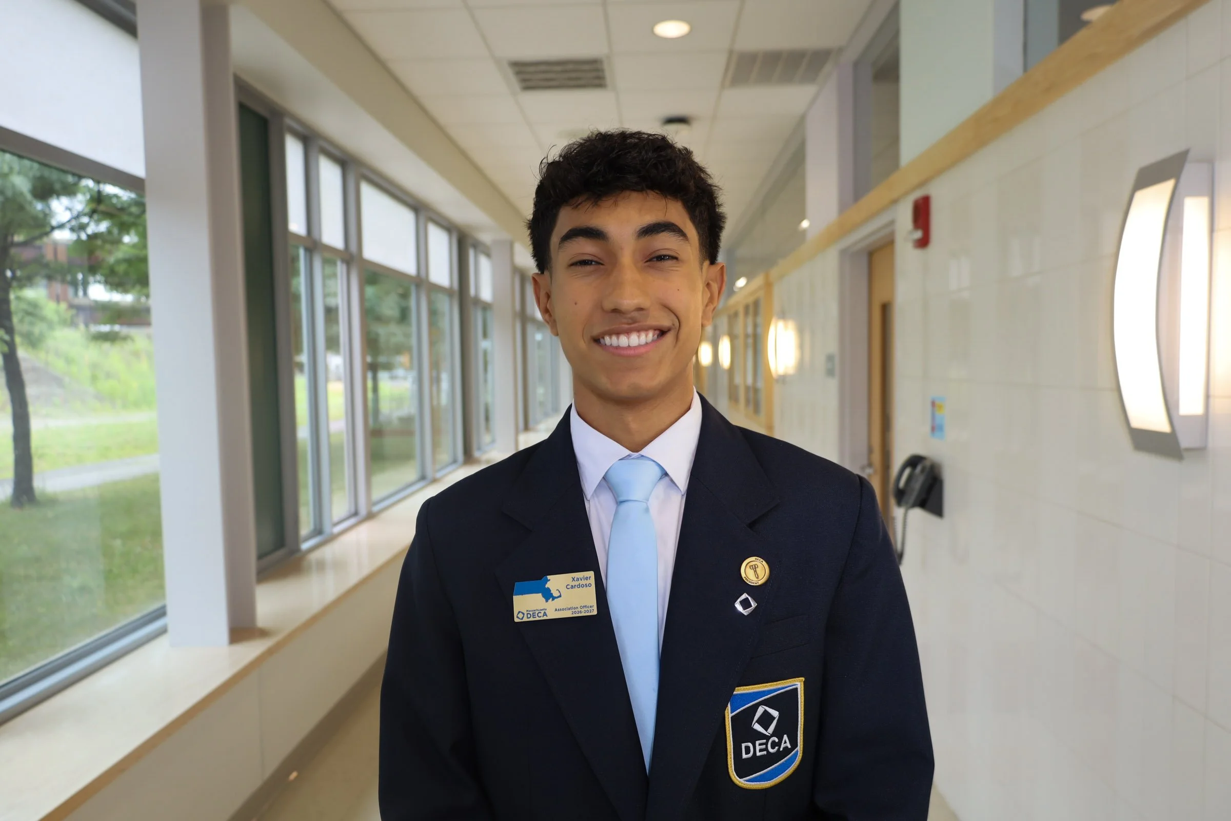 Young man in formal uniform smiling indoors near large windows with greenery outside.