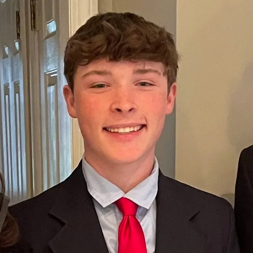 A young man with short, brown, wavy hair, smiling, wearing a black suit jacket, white dress shirt, and red tie, indoors near a window.
