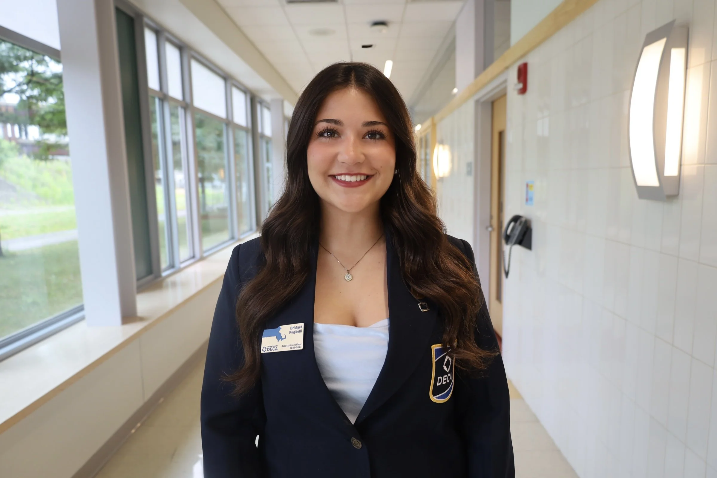 A young woman with long brown hair, wearing a black blazer with an association badge, is smiling while standing in a hallway with large windows and modern wall lights.