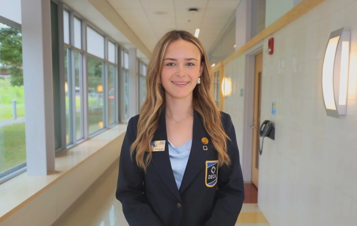 A young woman with long wavy blonde hair and a friendly smile, wearing a blue blazer with a DECA patch, standing in a well-lit hospital corridor with large windows and modern wall-mounted lights.