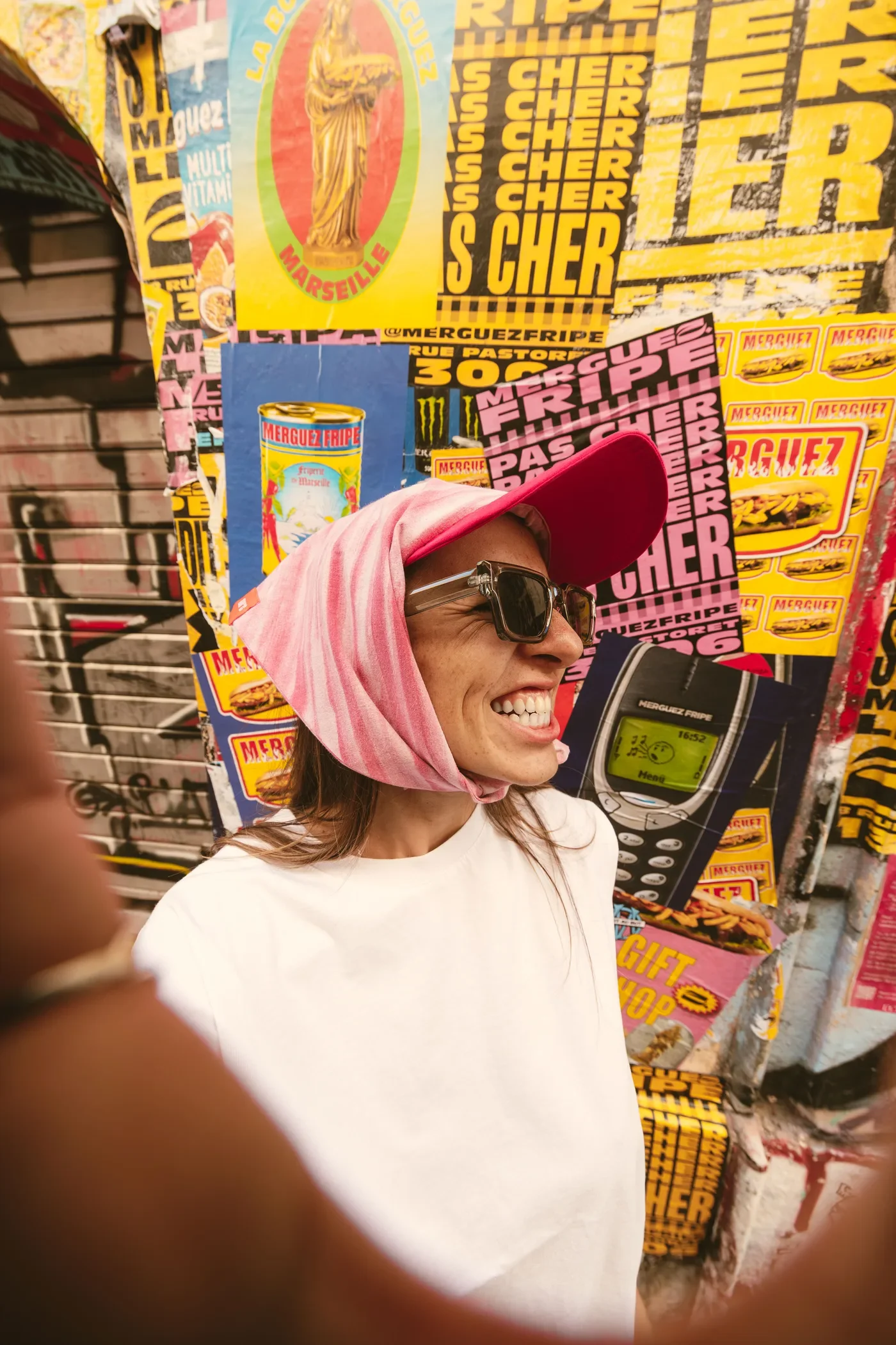 Une femme souriante portant une casquette rose, des lunettes de soleil foncées, une écharpe rose sur la tête et une chemise blanche, se tient devant un mur couvert d'affiches publicitaires colorées.