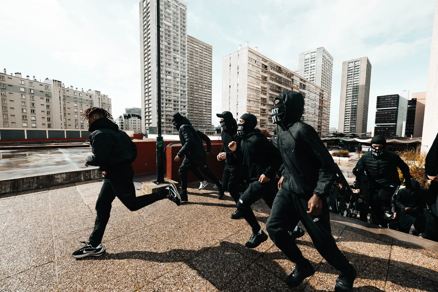 Groupes de personnes habillées en noir avec des masques, courant sur un toit urbain avec des immeubles modernes en arrière-plan.