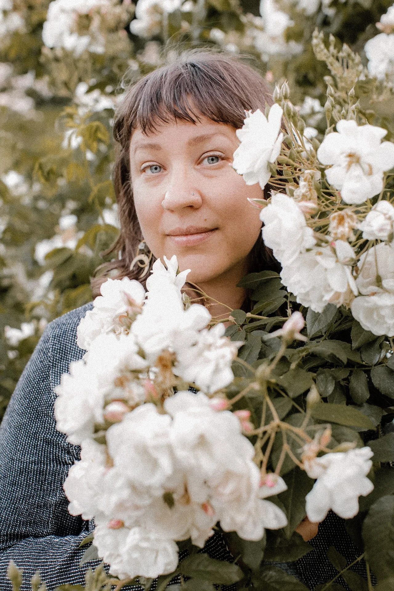 A woman with short dark hair and light blue eyes is surrounded by white flowers and leaves, outdoors.