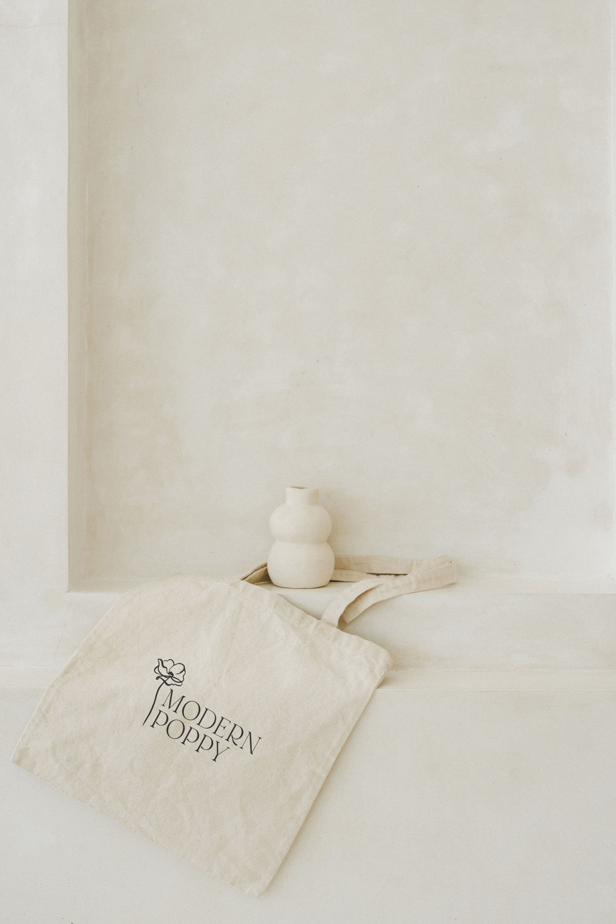Cream-colored tote bag with 'MODERN POPPY' logo and floral design, placed on a white surface with a ceramic vase in the background.