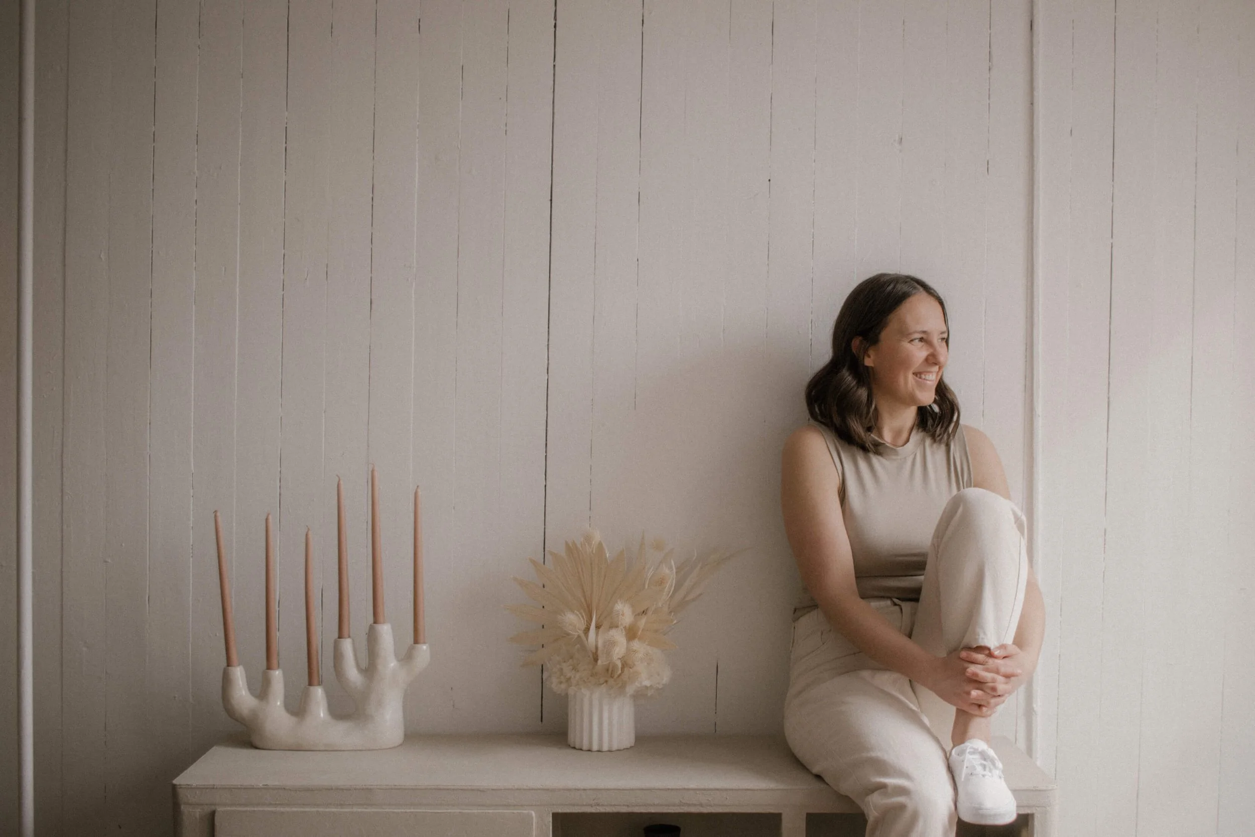Woman seated on a bench with decor, including a hand-shaped candle holder and dried floral arrangement, against a light wooden wall.