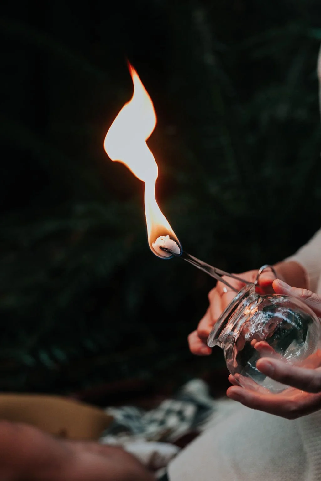 Person holding a glass jar with a lit match or similar stick, creating a flame in a dark outdoor setting.