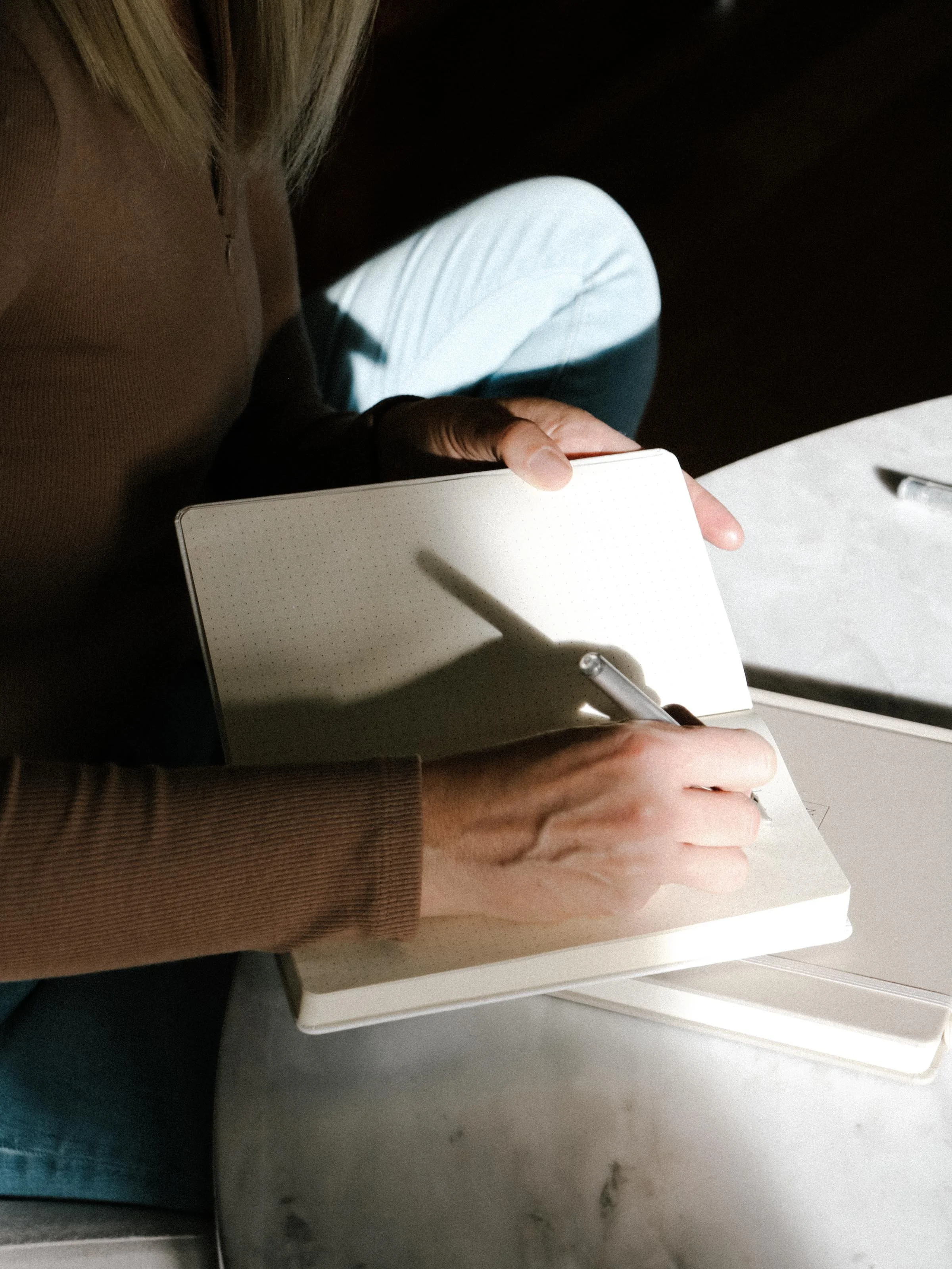 A person sitting at a table, writing in a notebook with a pen. The person is wearing a brown long sleeve shirt and white pants. The shadow of the pen is cast on the open page.