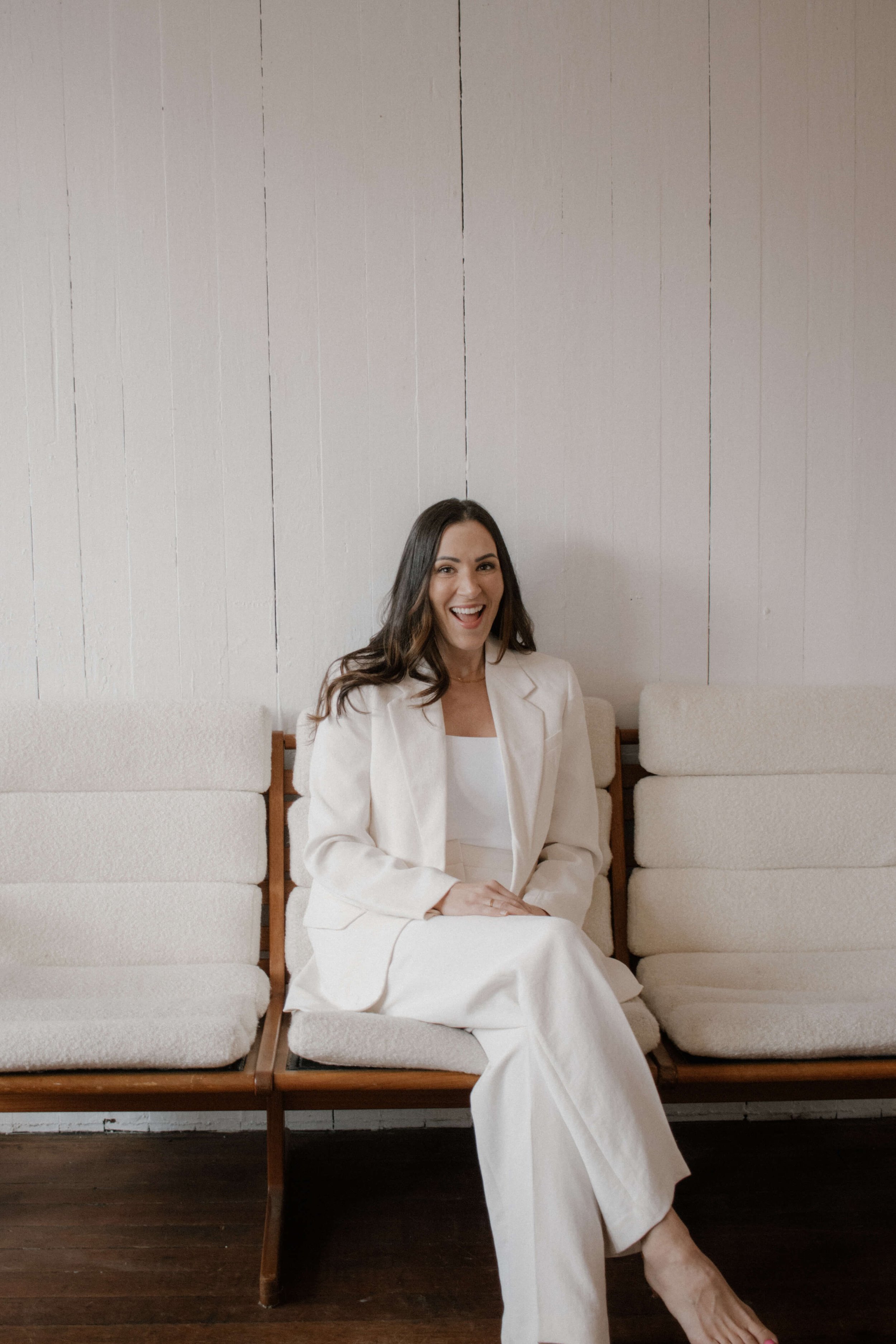 Woman in a white suit sitting on a wooden bench, smiling and looking at the camera, with a light-colored wall behind her.