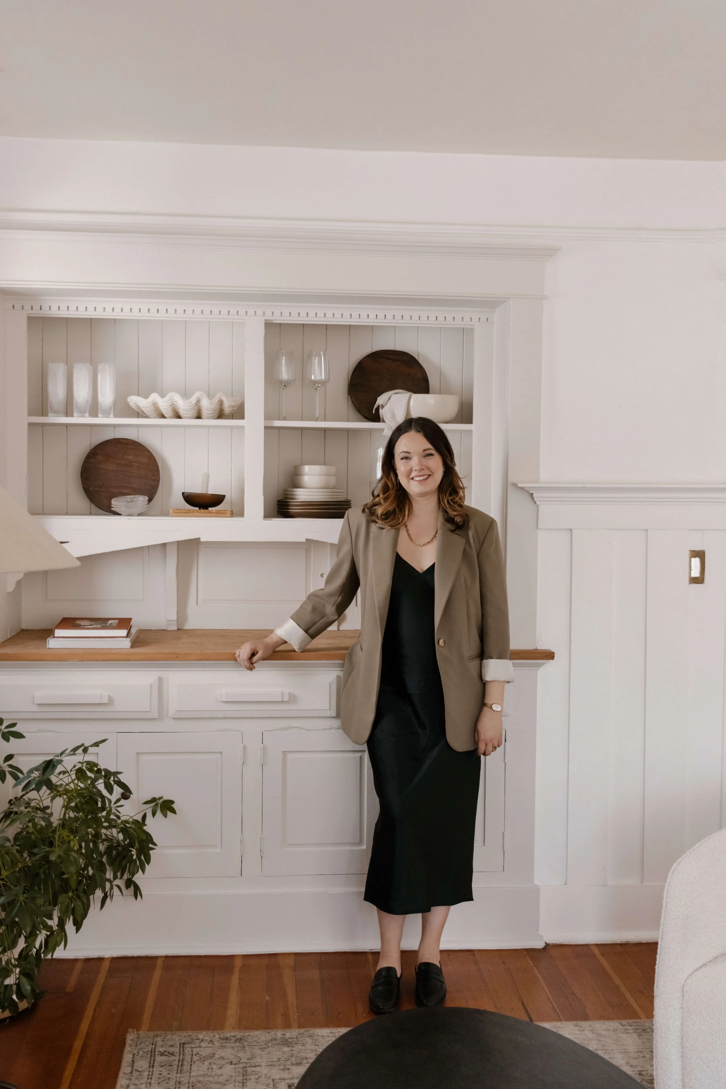 A woman standing in a dining room, leaning against a built-in white wooden cabinet with open shelves, dressed in a beige blazer, black dress, black loafers, smiling at the camera.