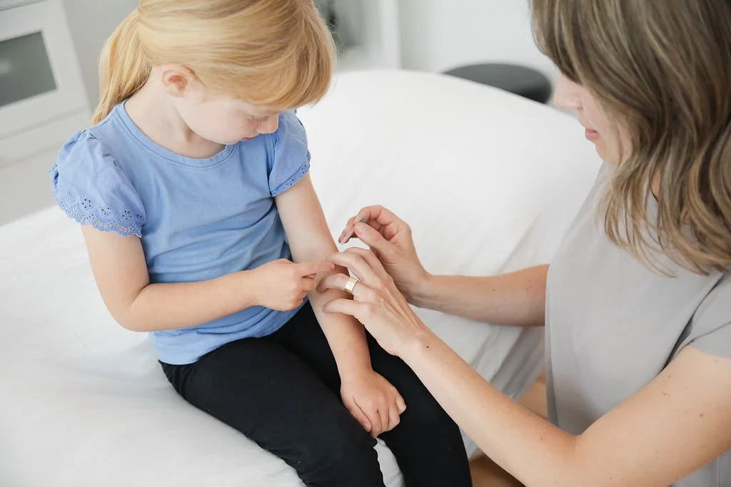 A young girl sitting on a bed receiving a vaccination from a healthcare worker in a clinical setting.