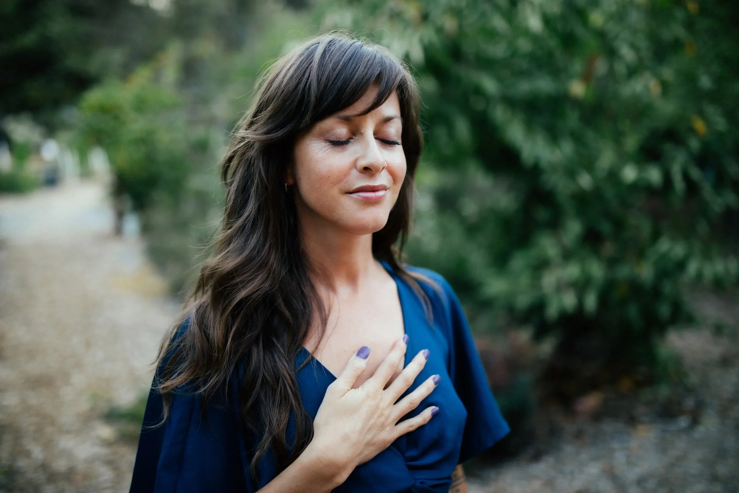 A woman with long dark hair and a nose ring wearing a blue top standing outdoors with her eyes closed, hand on her chest, appearing peaceful.