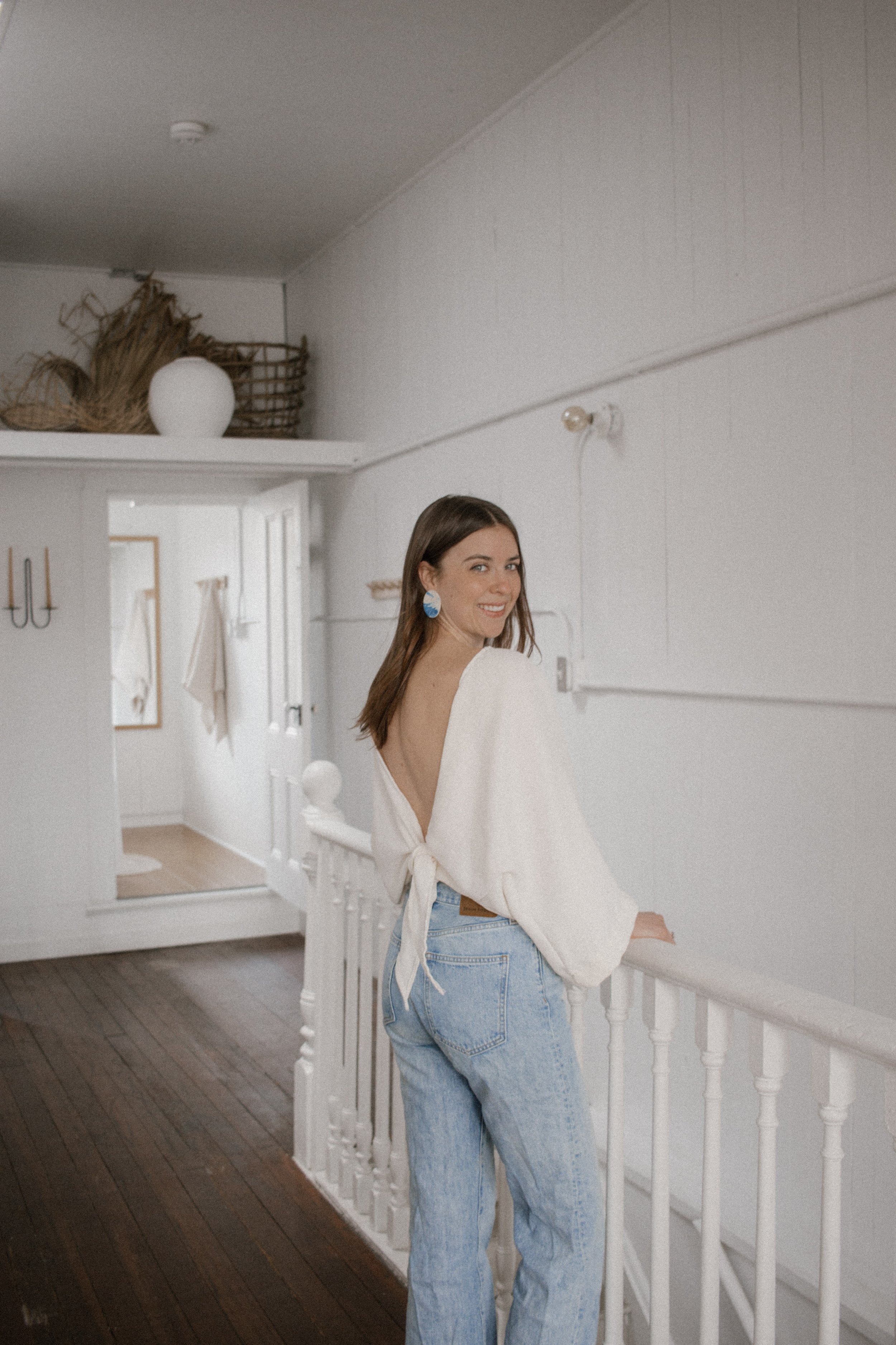 Woman in an open-back top and jeans standing in a white interior space, smiling at the camera.