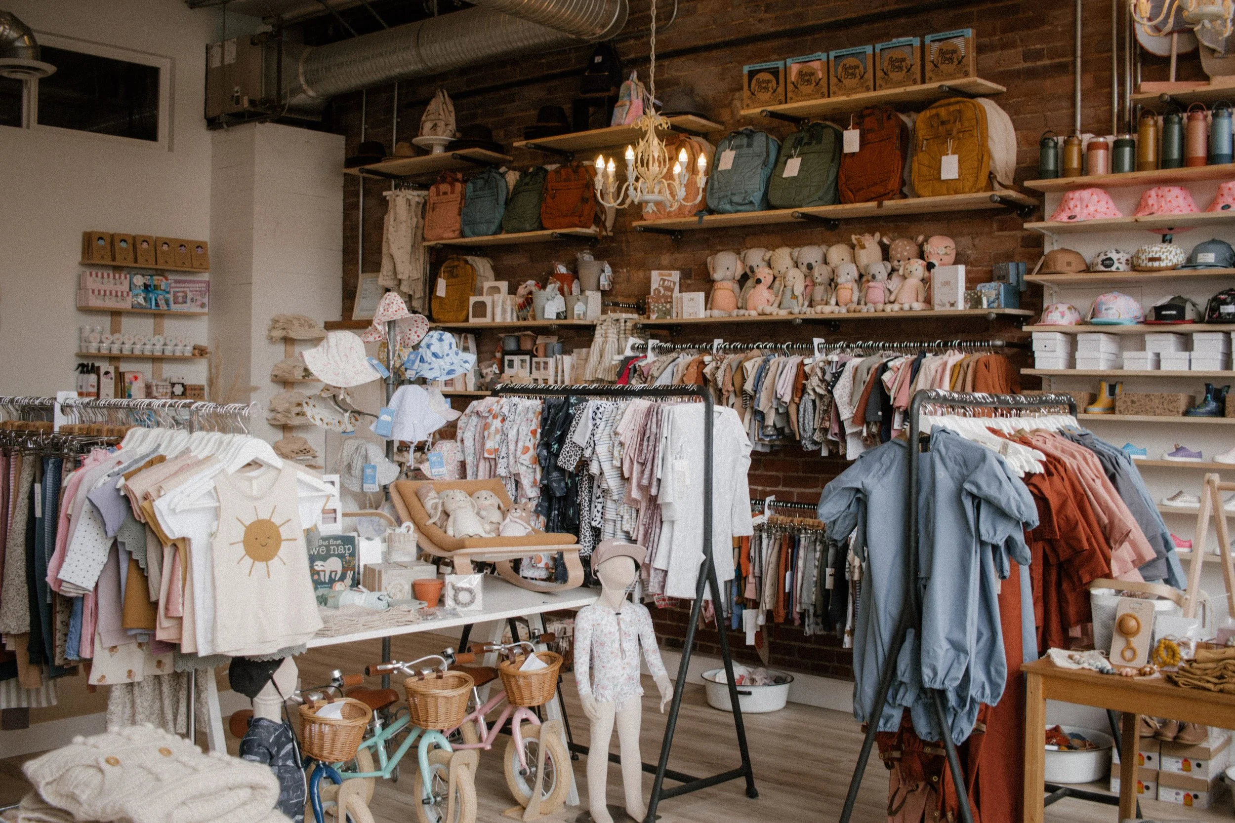 Interior of a children's clothing and toy store with shelves stocked with toys, clothing, backpacks, hats, and accessories, and clothing racks displaying various children's garments.