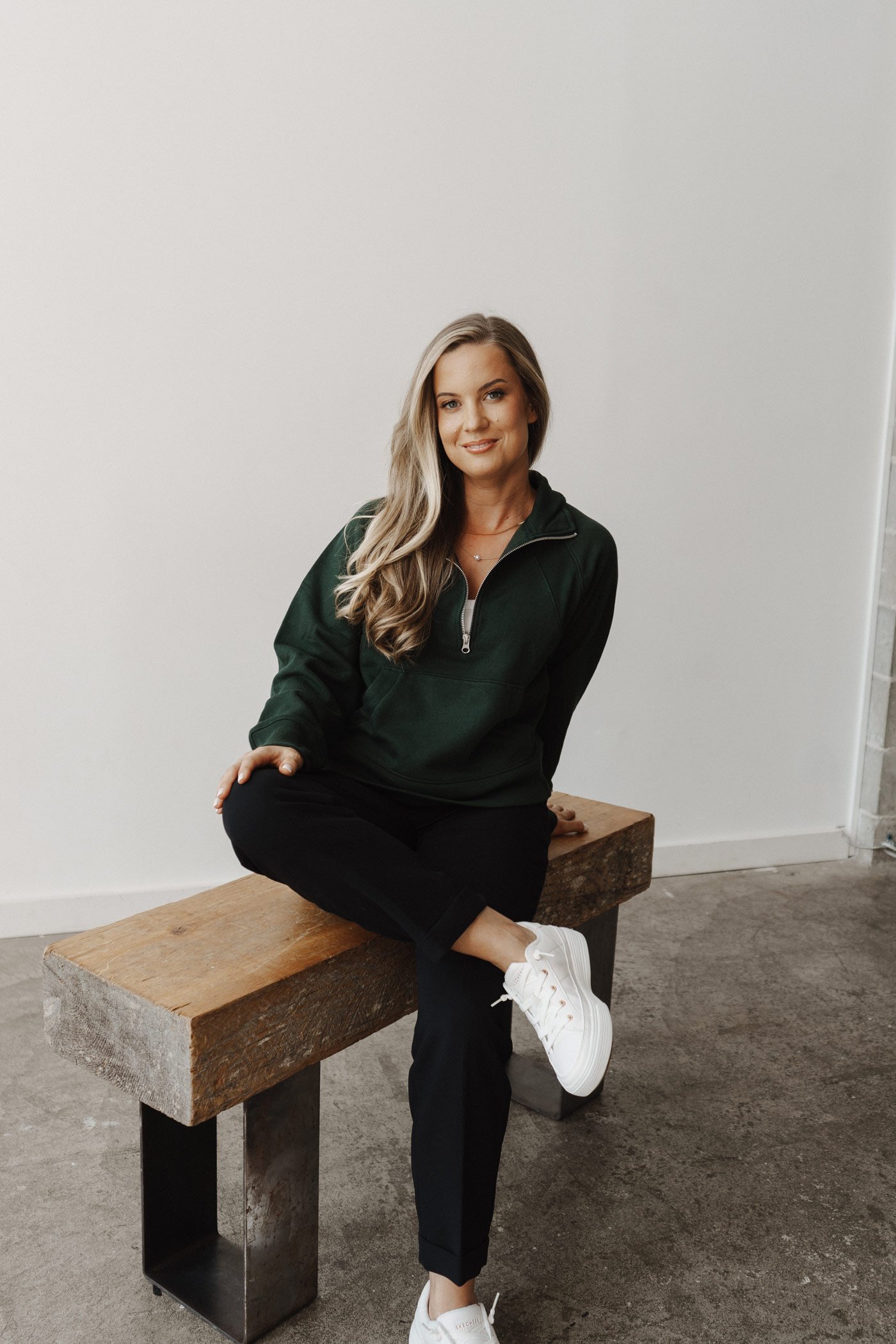 A young woman with long, wavy blonde hair sitting on a wooden bench against a plain white wall. She is dressed in a dark green zip-up jacket, black pants, and white sneakers. She is smiling at the camera with her right ankle resting on her left knee.