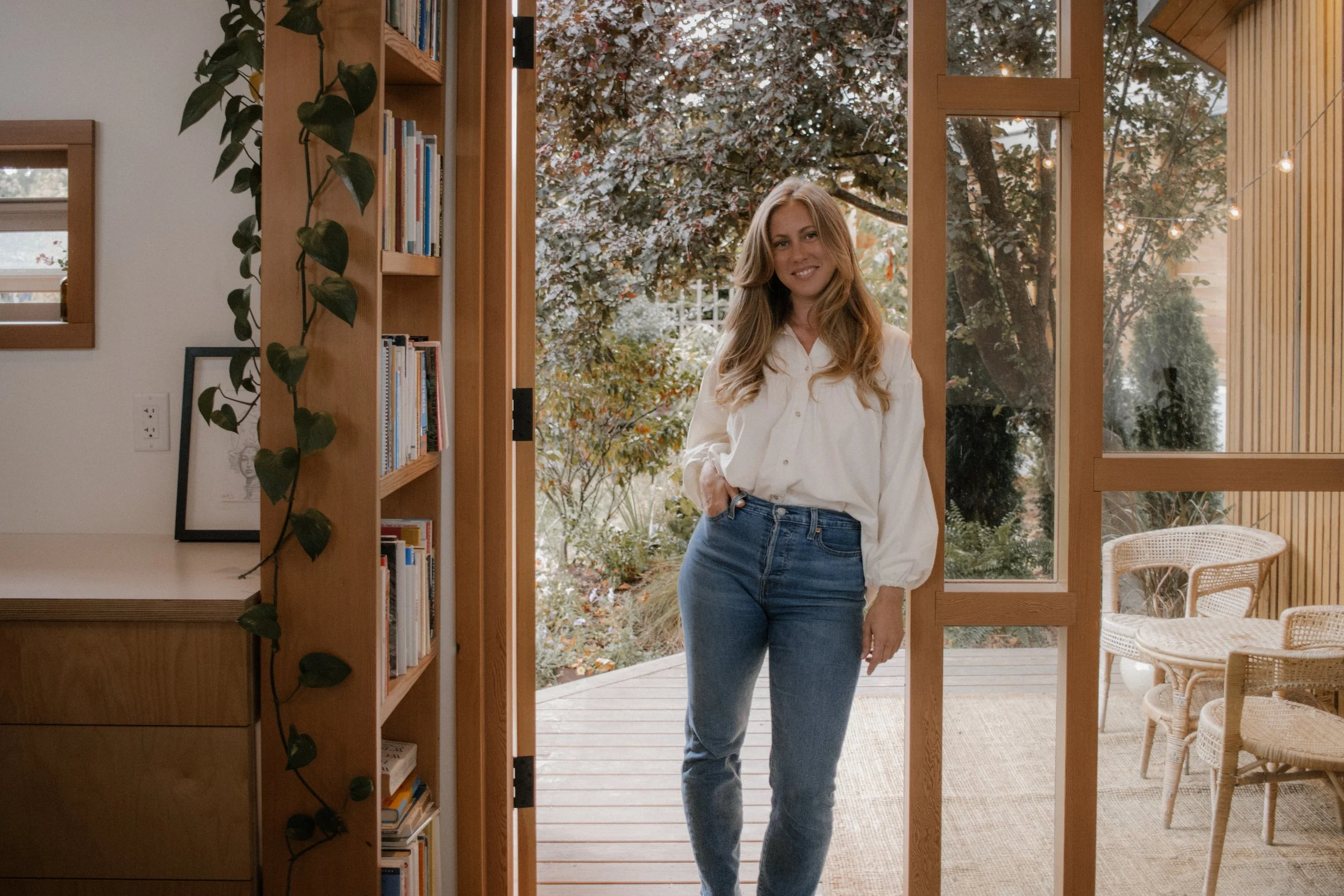 Woman standing in a doorway next to a bookshelf, wearing a white blouse and jeans, with a garden in the background.