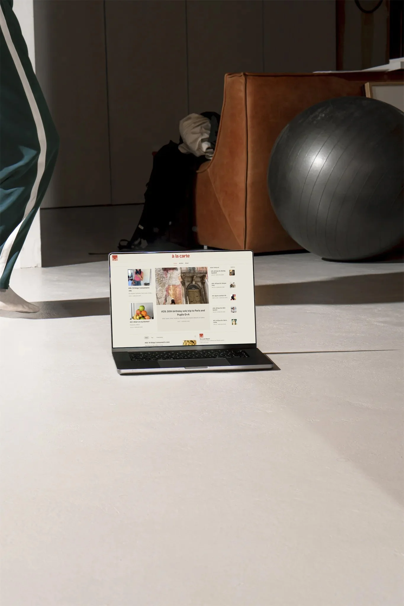 Laptop on a light-colored floor displaying a website, in a room with a brown couch, a large black exercise ball, a stool with a green and white striped jacket, and a bag with clothes.