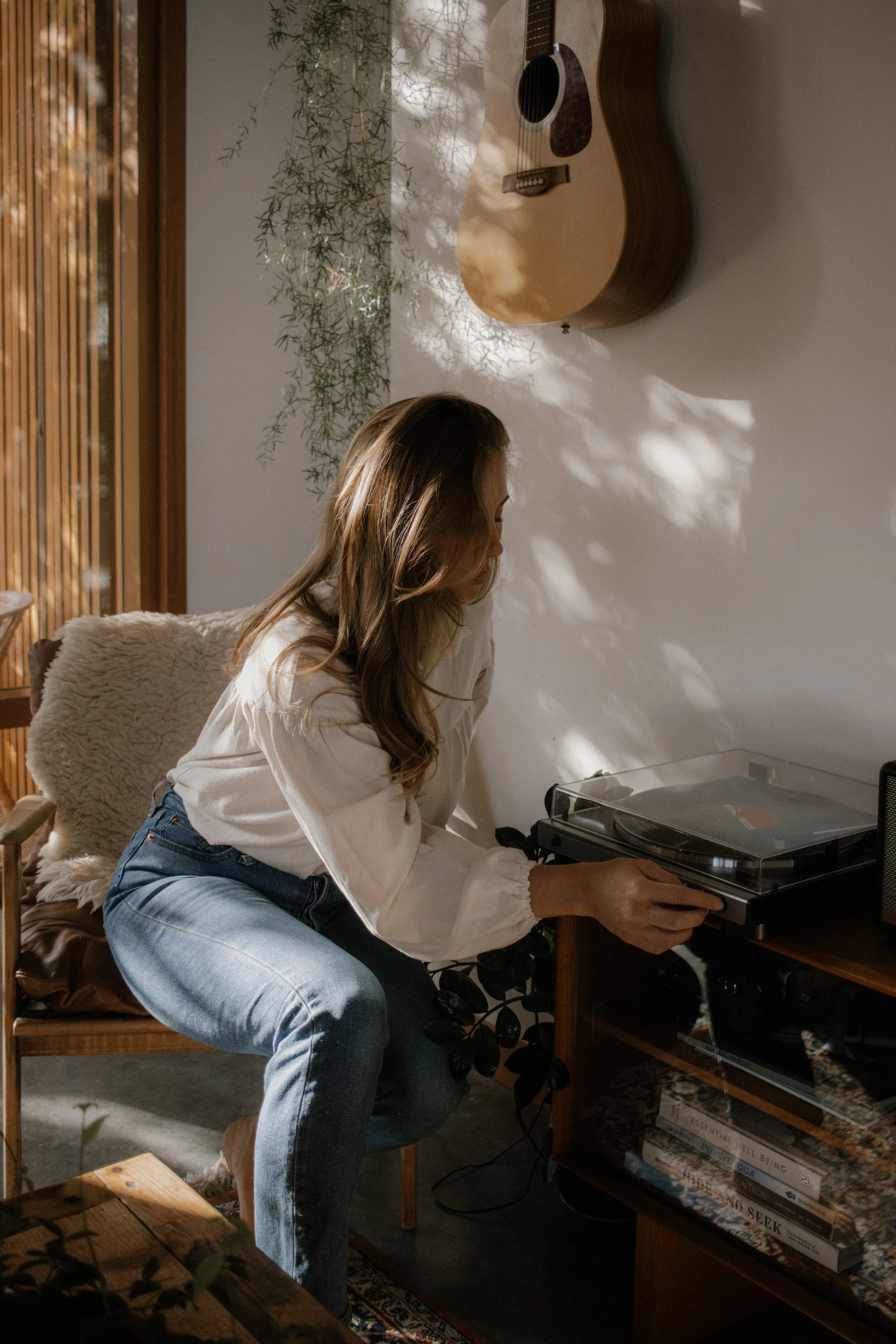 Woman adjusting a record player in a cozy room with a guitar on the wall and plants nearby.