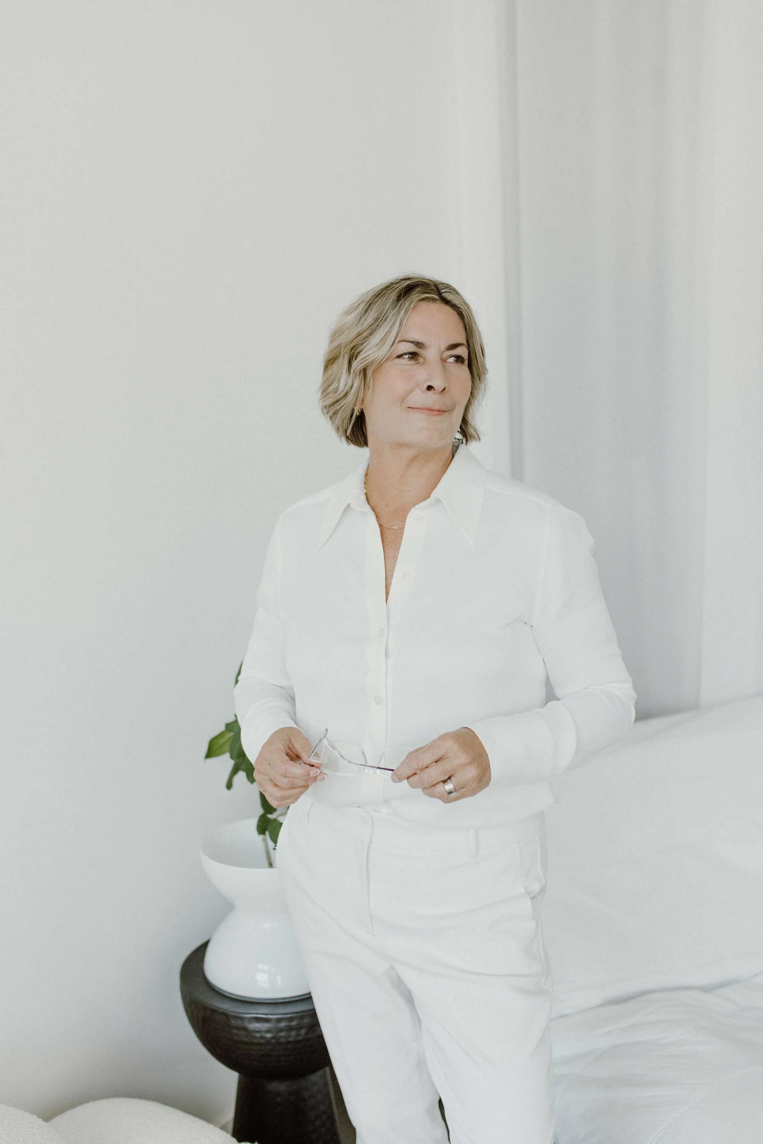 A woman with short blonde hair wearing a white shirt and pants, holding glasses, standing in a minimalistic, white room.
