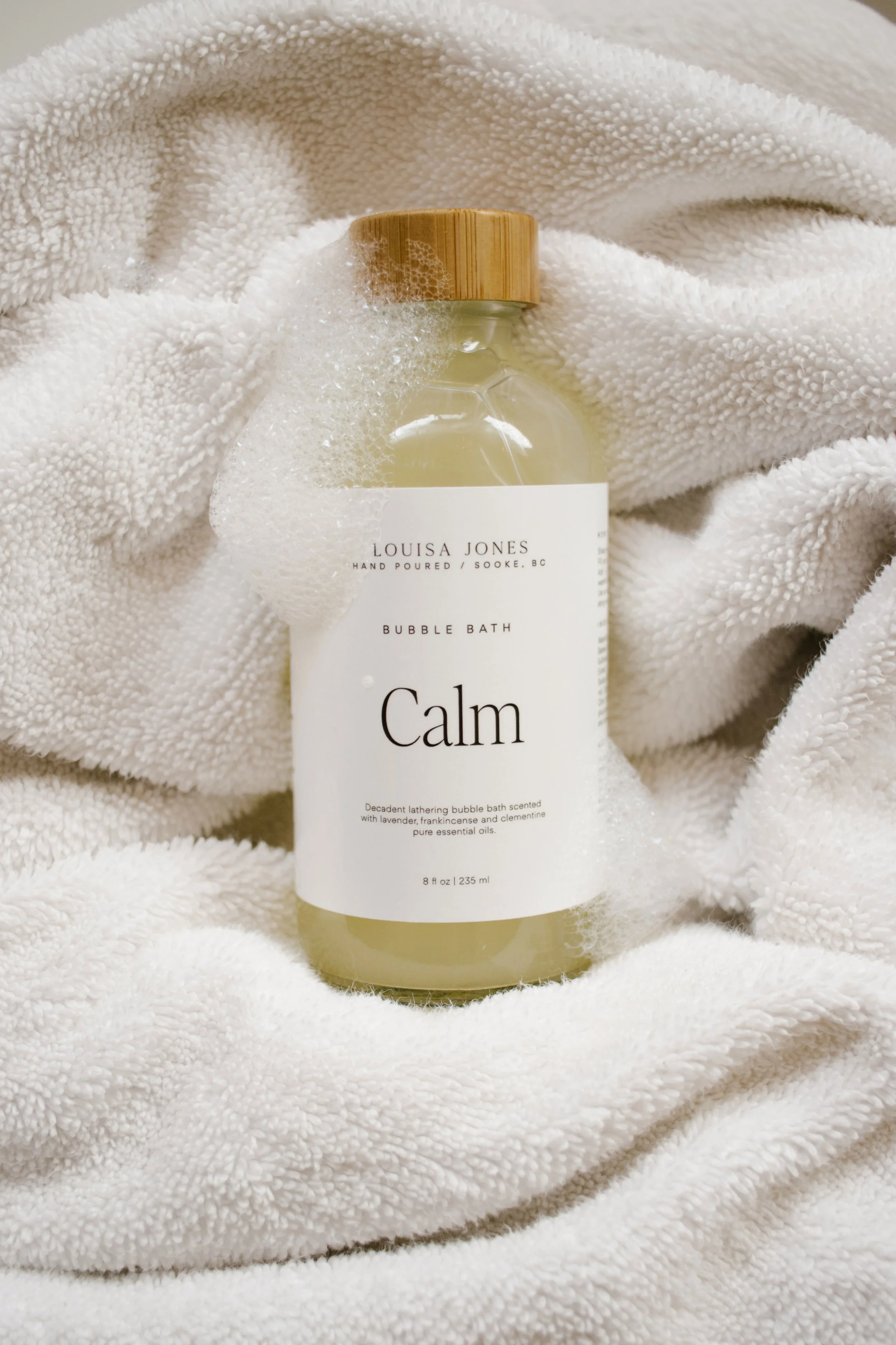 A bottle of Louisa Jones Calming Bubble Bath with a bamboo cap, nestled in soft white towels.