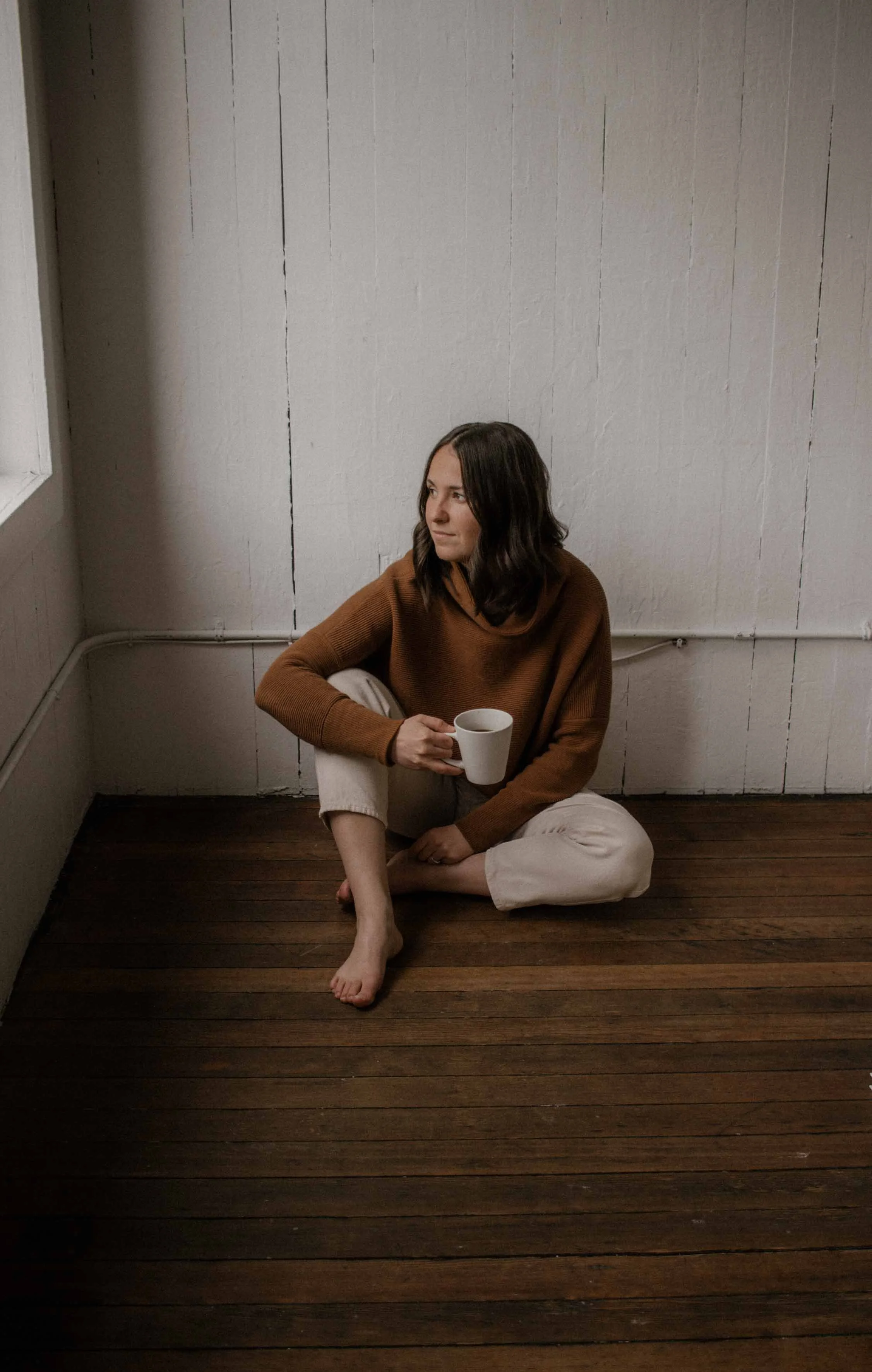 Woman sitting on wooden floor holding a mug, wearing a brown sweater and light pants, looking out a window in a white-walled room.