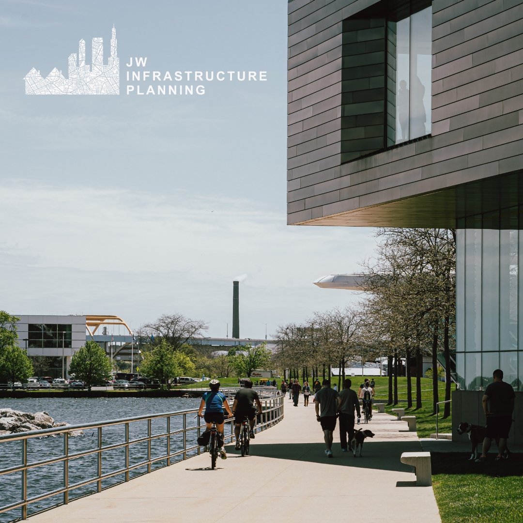People walking and biking along a riverside walkway near modern buildings with trees and industrial smokestack in the background.