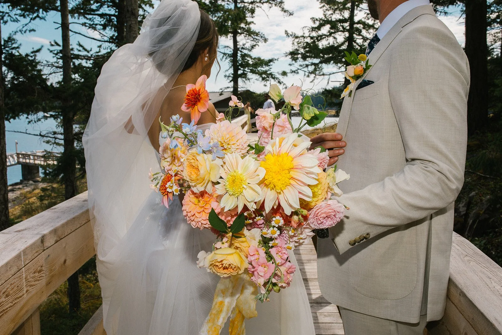 Bride and groom exchanging wedding vows outdoors, with the bride holding a large bouquet of colorful flowers, and trees and water in the background.