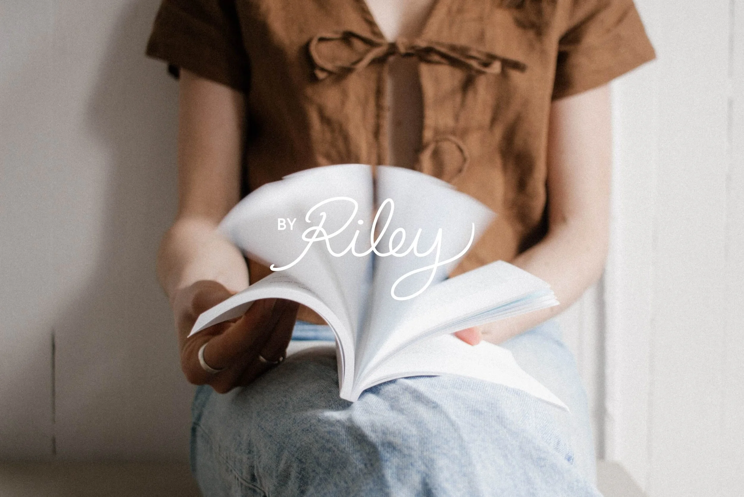 Person reading a magazine or book while sitting down, wearing a brown top with a bow detail, and blue jeans, against a white wall.